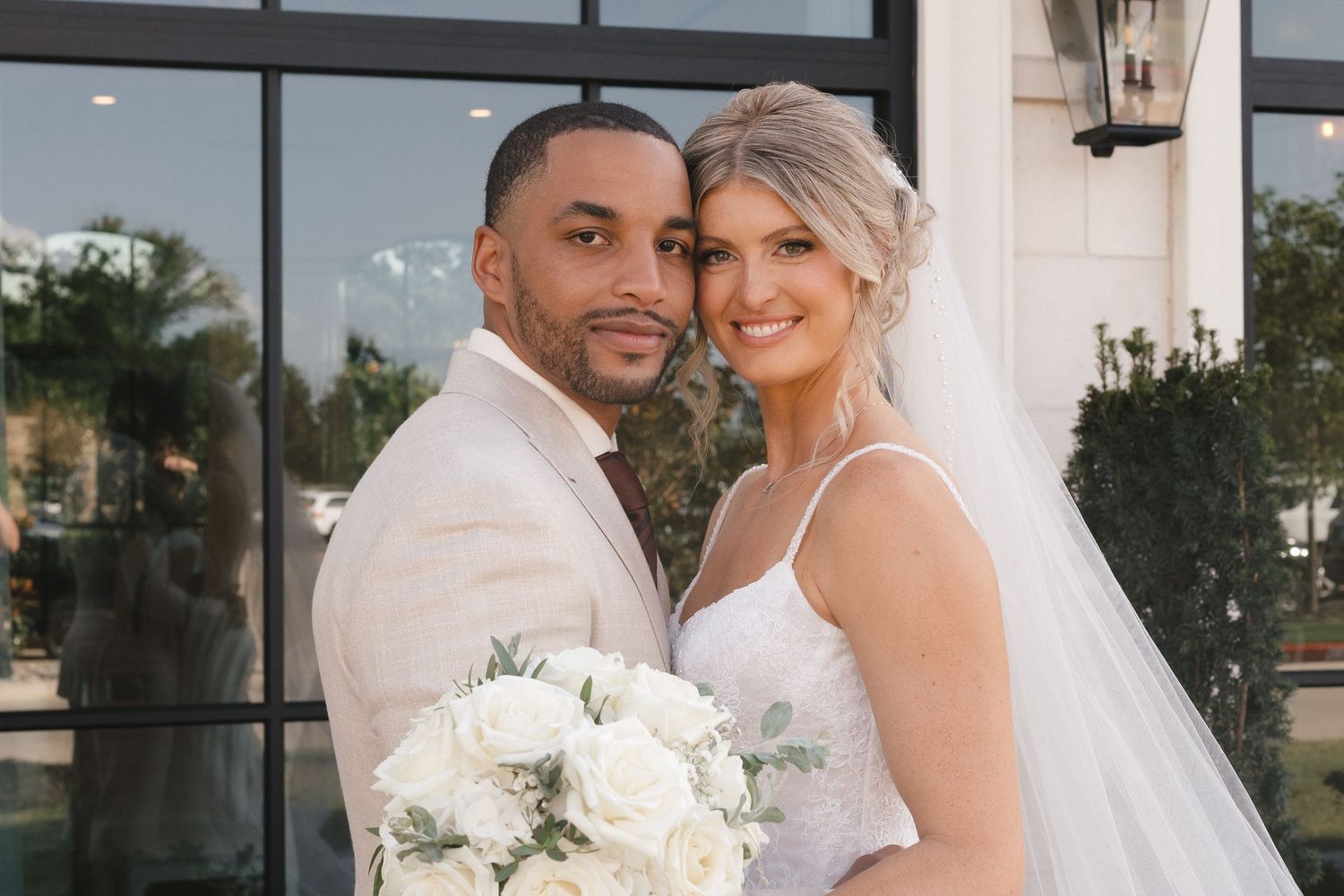 A couple poses in wedding attire, groom in beige suit, bride in white gown holding bouquet, smiling outdoors.
