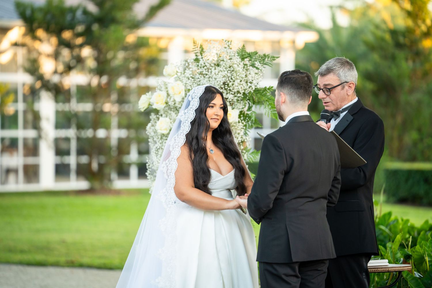 Bride and groom exchanging vows outdoors with officiant, floral arch backdrop, lush greenery, elegant wedding ceremony.
