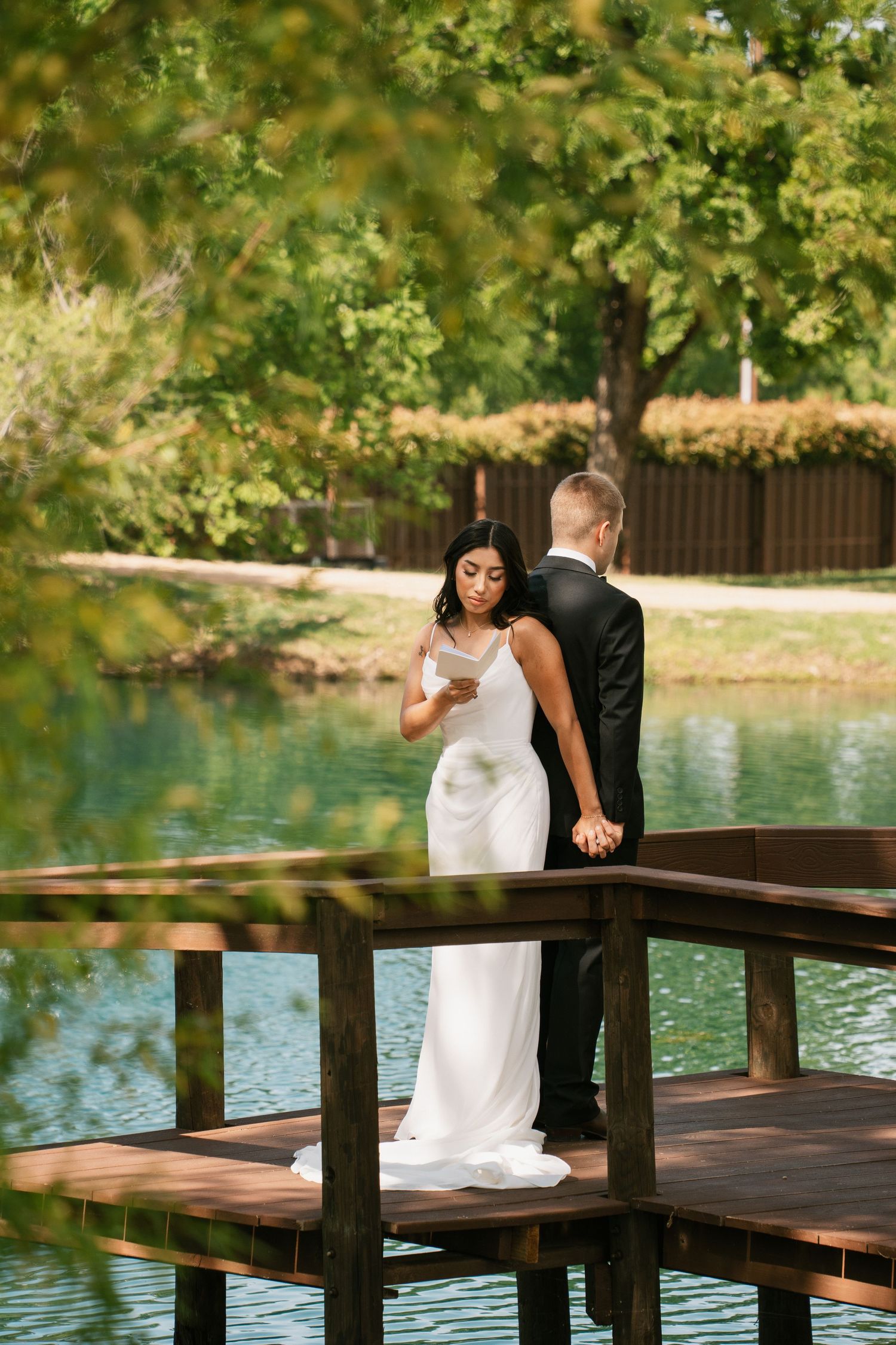 Bride and groom holding hands on wooden dock by serene lake, surrounded by lush green trees on their wedding day.
