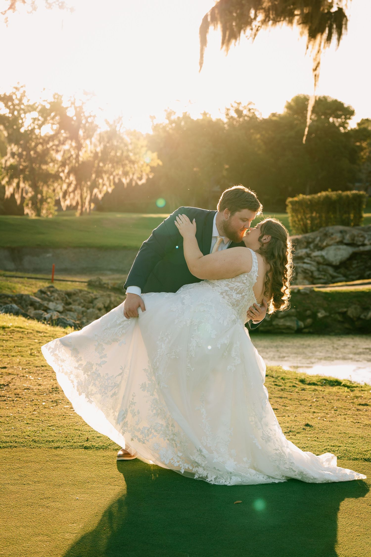 Groom dips bride in lace wedding gown at golden hour on golf course, sharing romantic kiss with sunlit trees behind.