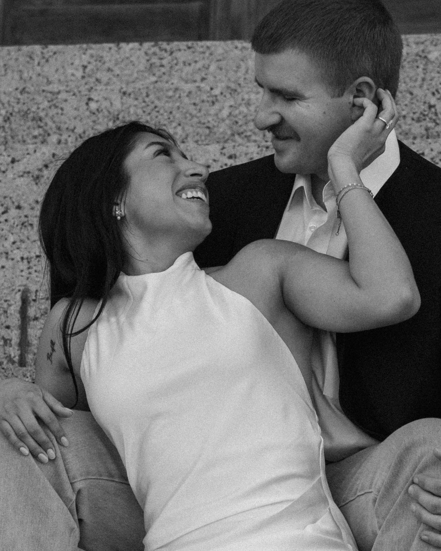 Black and white photo of laughing couple gazing at each other while relaxing against a stone wall.