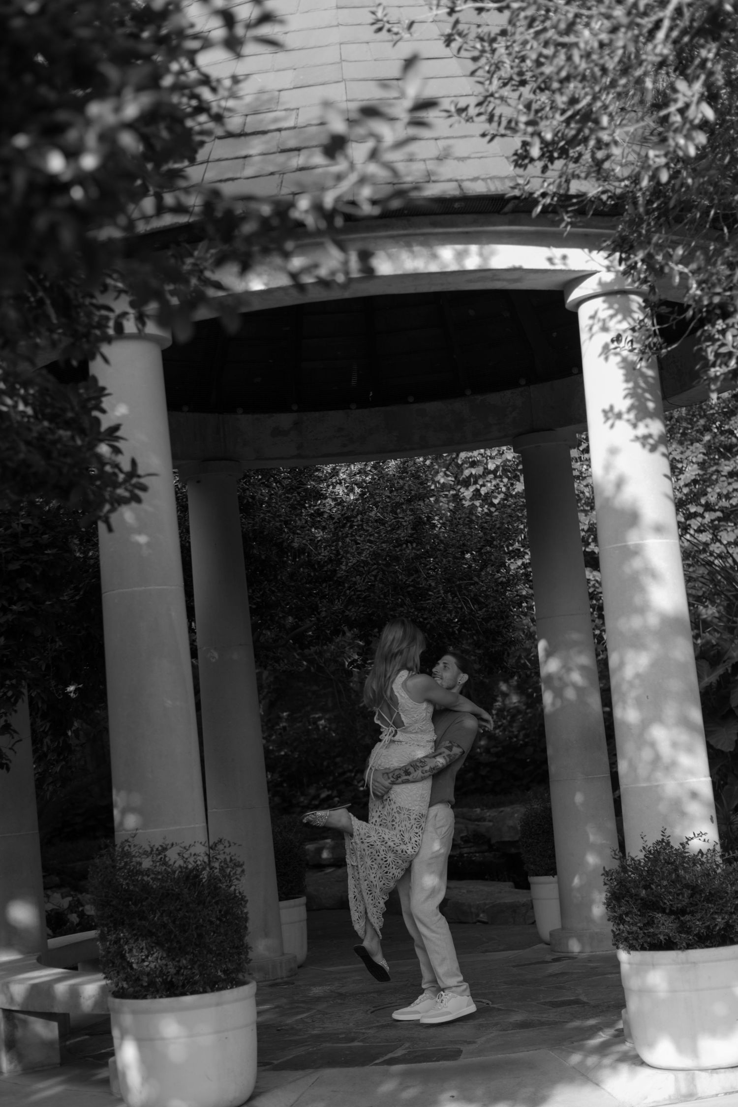 Black and white photo of a couple embracing as he lifts her beneath a classic columned garden gazebo surrounded by trees and potted shrubs.