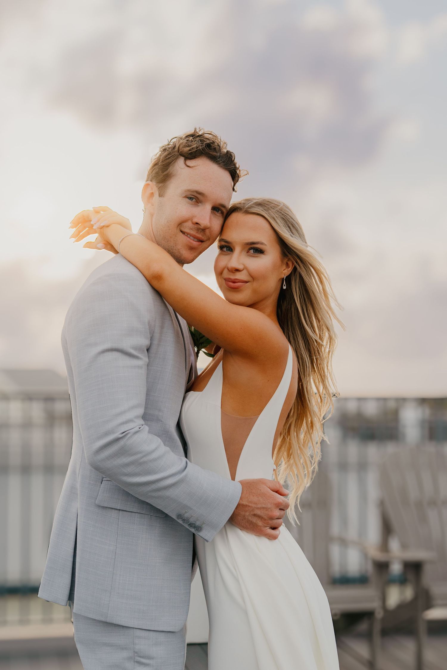 Bride and groom embrace at golden sunset on waterfront deck, she wears deep V-neck wedding gown.