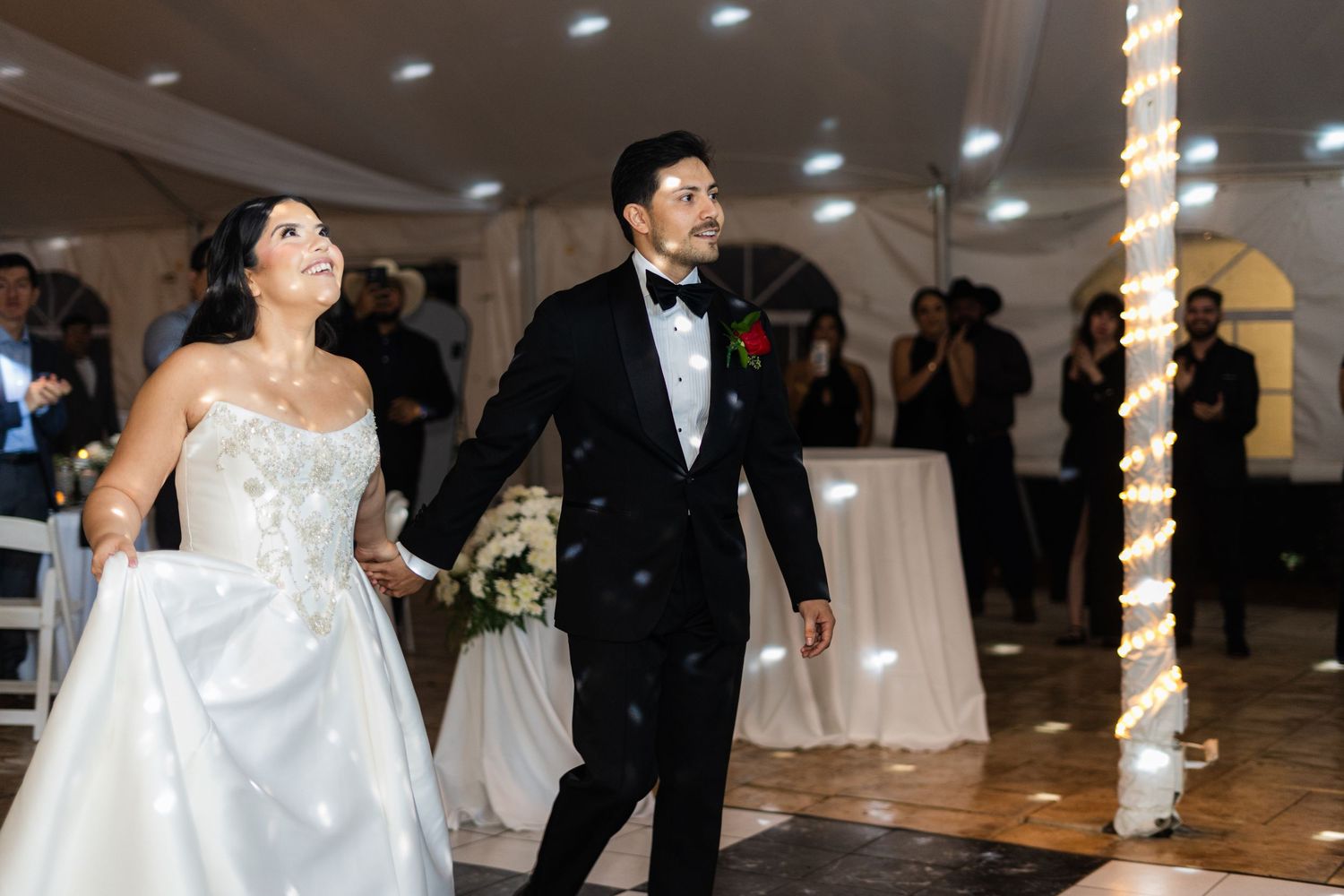 Bride in strapless gown and groom in tuxedo share first dance at elegant wedding reception under white tent.