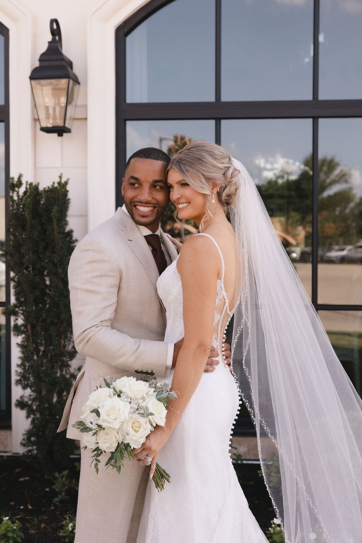 A smiling bride and groom pose outdoors, she holds white roses, wearing a fitted gown with veil, he wears a beige suit.
