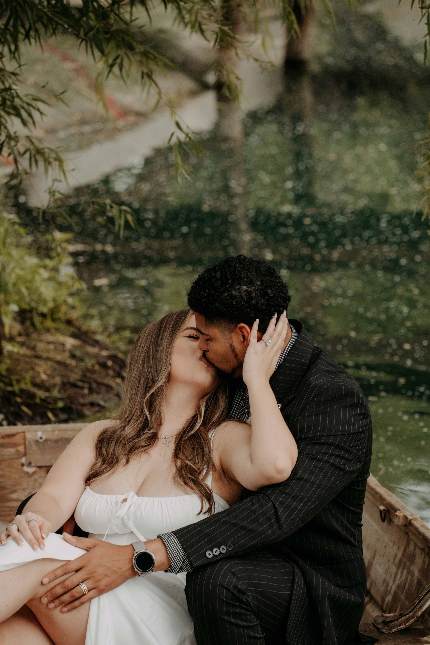 Couple sharing a tender kiss in a wooden boat surrounded by lush greenery and water reflections.