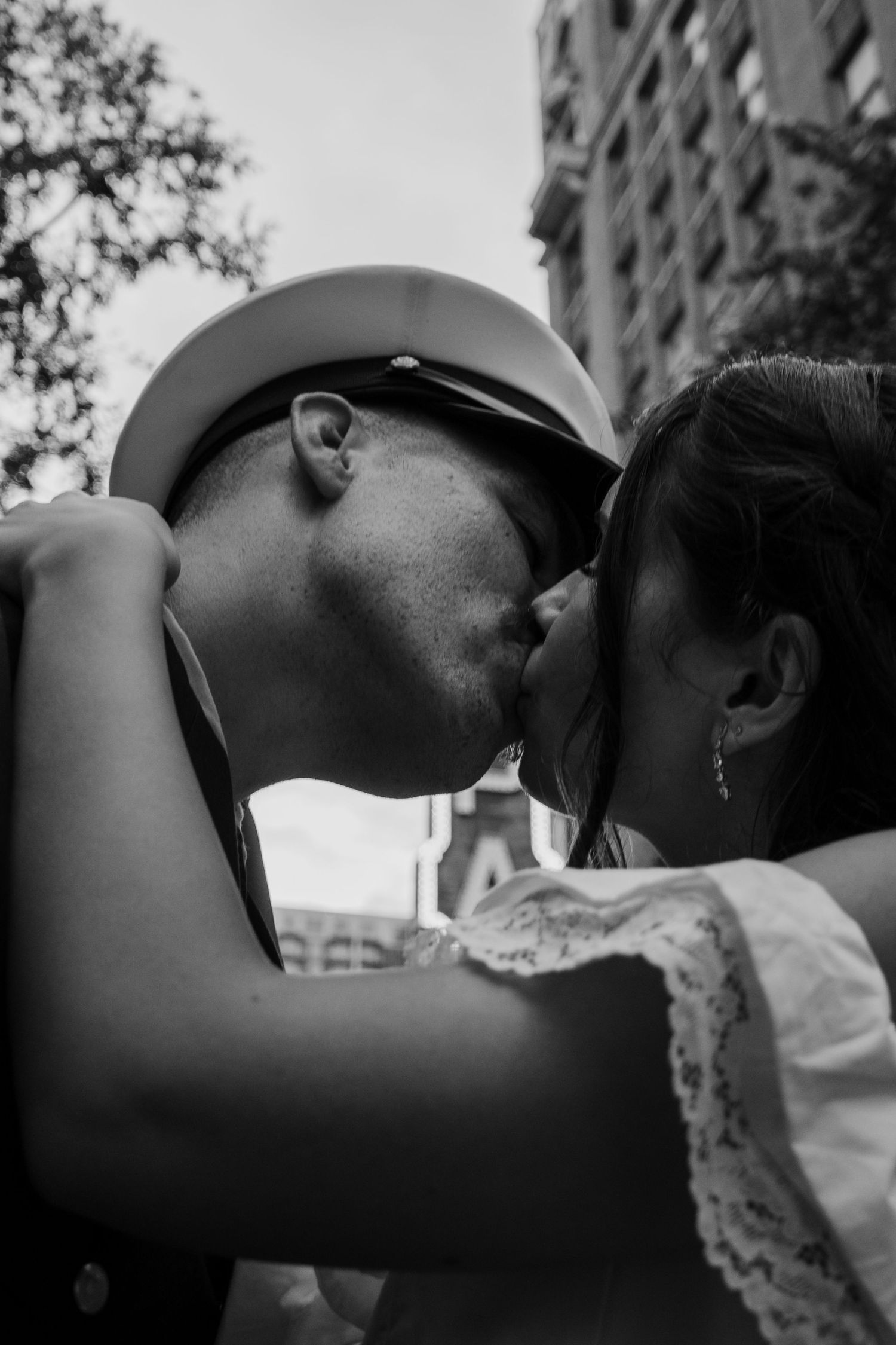 Black and white photo of a couple sharing a romantic kiss outdoors, man wearing a cap and sunglasses.