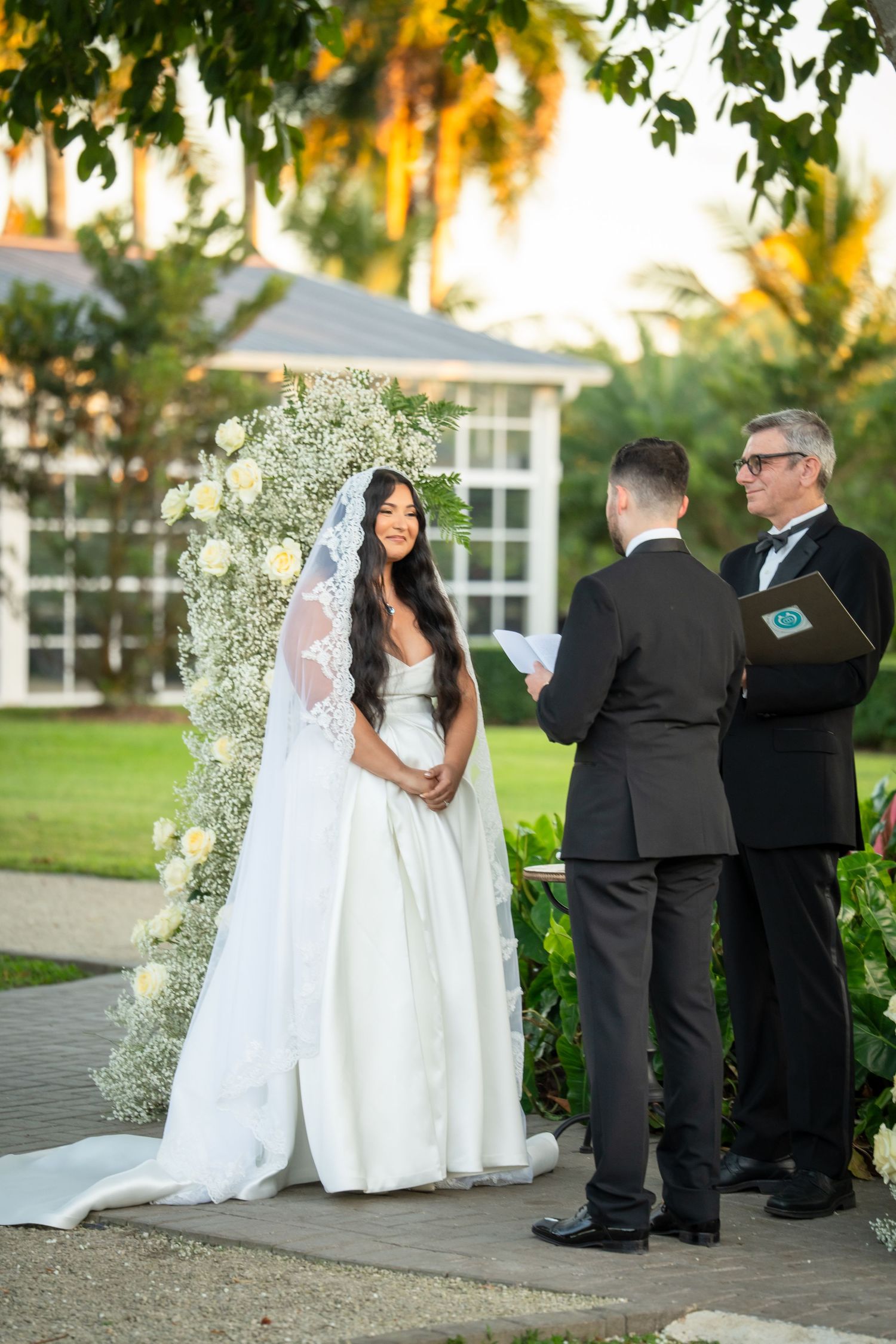 Bride smiling at groom during outdoor wedding ceremony with white floral arch and baby's breath backdrop.
