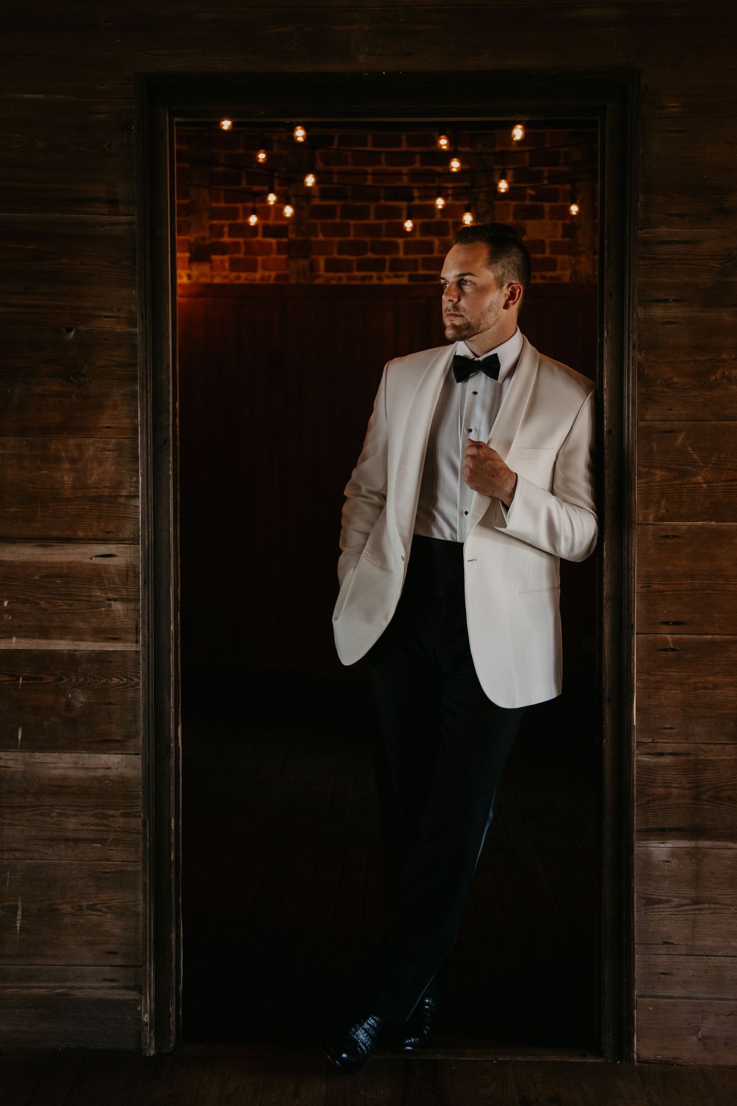 Groom in white tuxedo jacket and black bow tie standing in a doorway with warm string lights on brick wall.