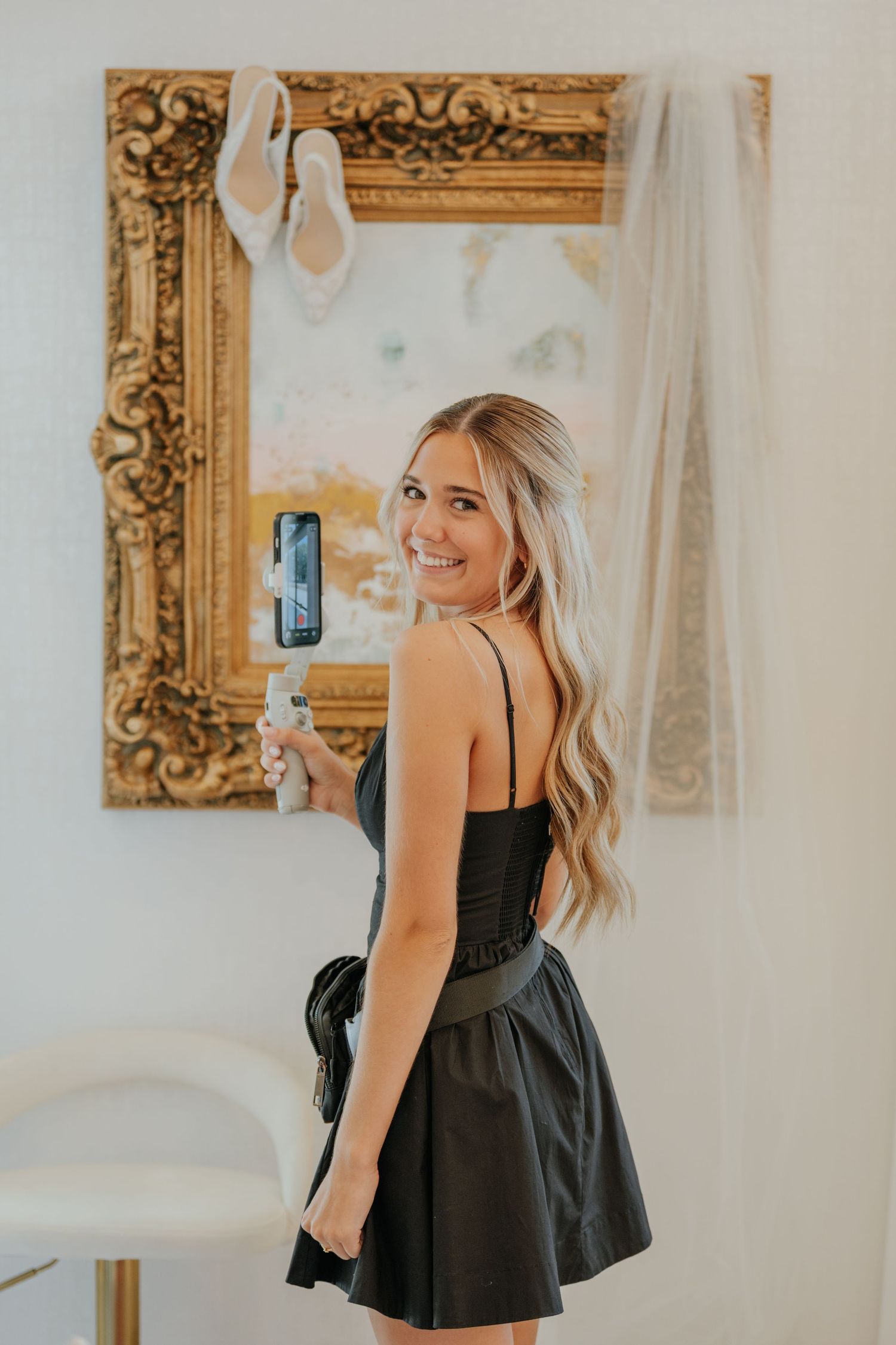 Smiling blonde woman in a black dress holding a phone in front of an ornate gold mirror.