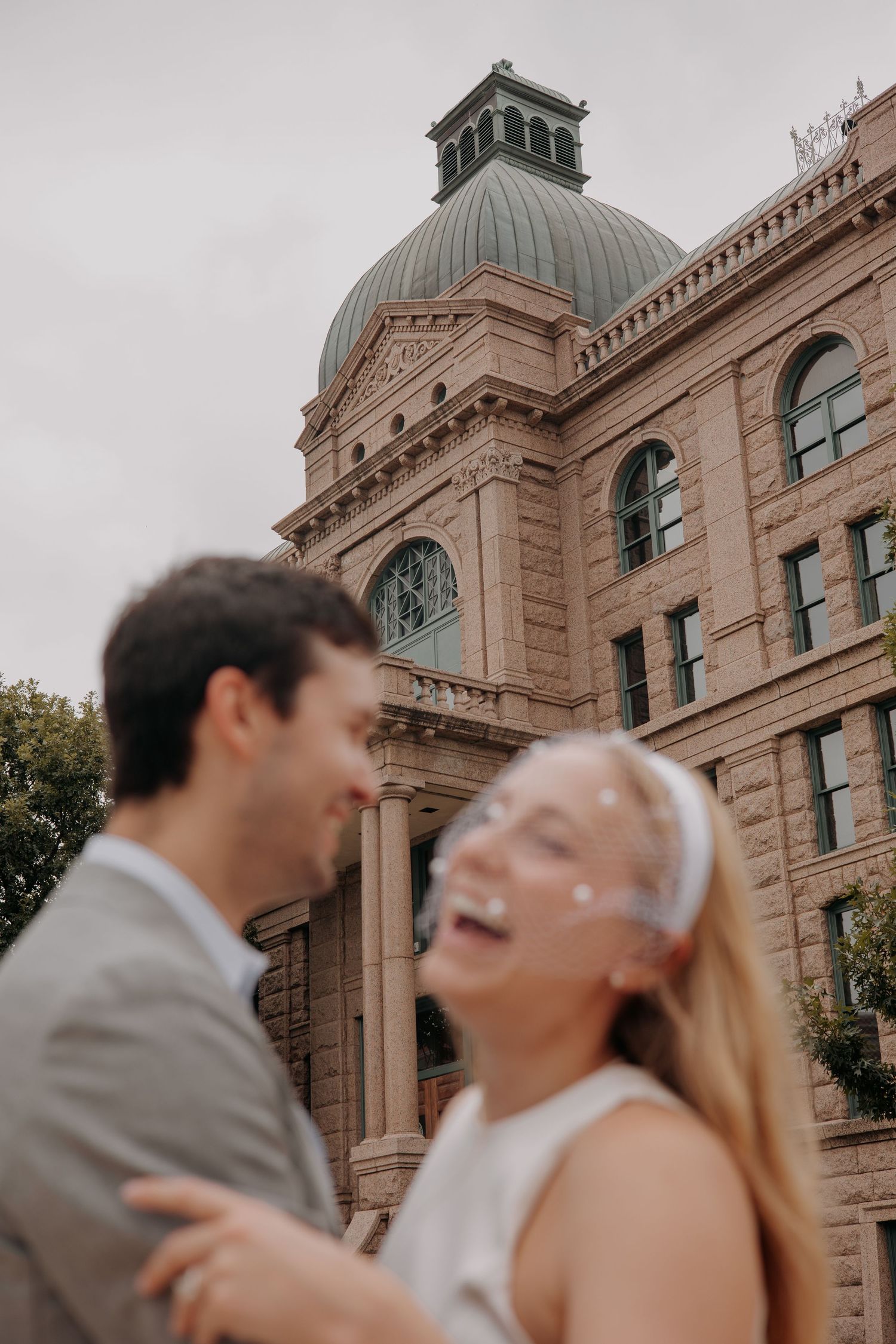 Smiling couple embracing in front of a historic sandstone courthouse with a green domed roof, perfect for wedding or engagement photos.