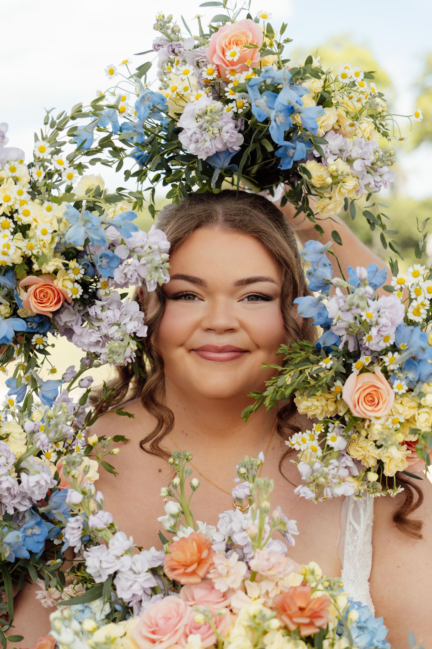 Smiling woman surrounded by a large floral arch with blue, peach, and white flowers, wearing a flower crown outdoors.