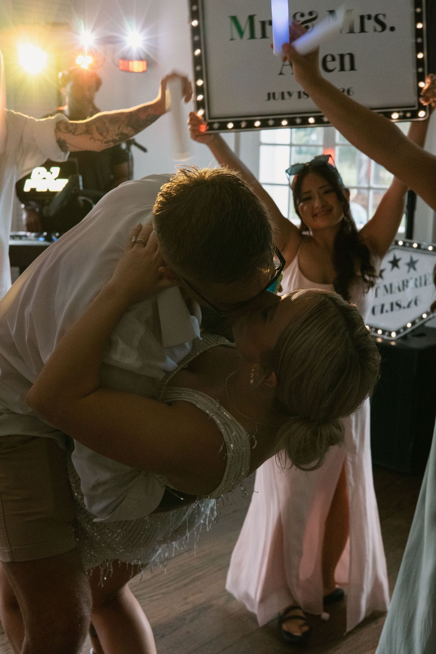 Newlyweds share a romantic dip kiss on the dance floor at their wedding reception with guests celebrating behind them.