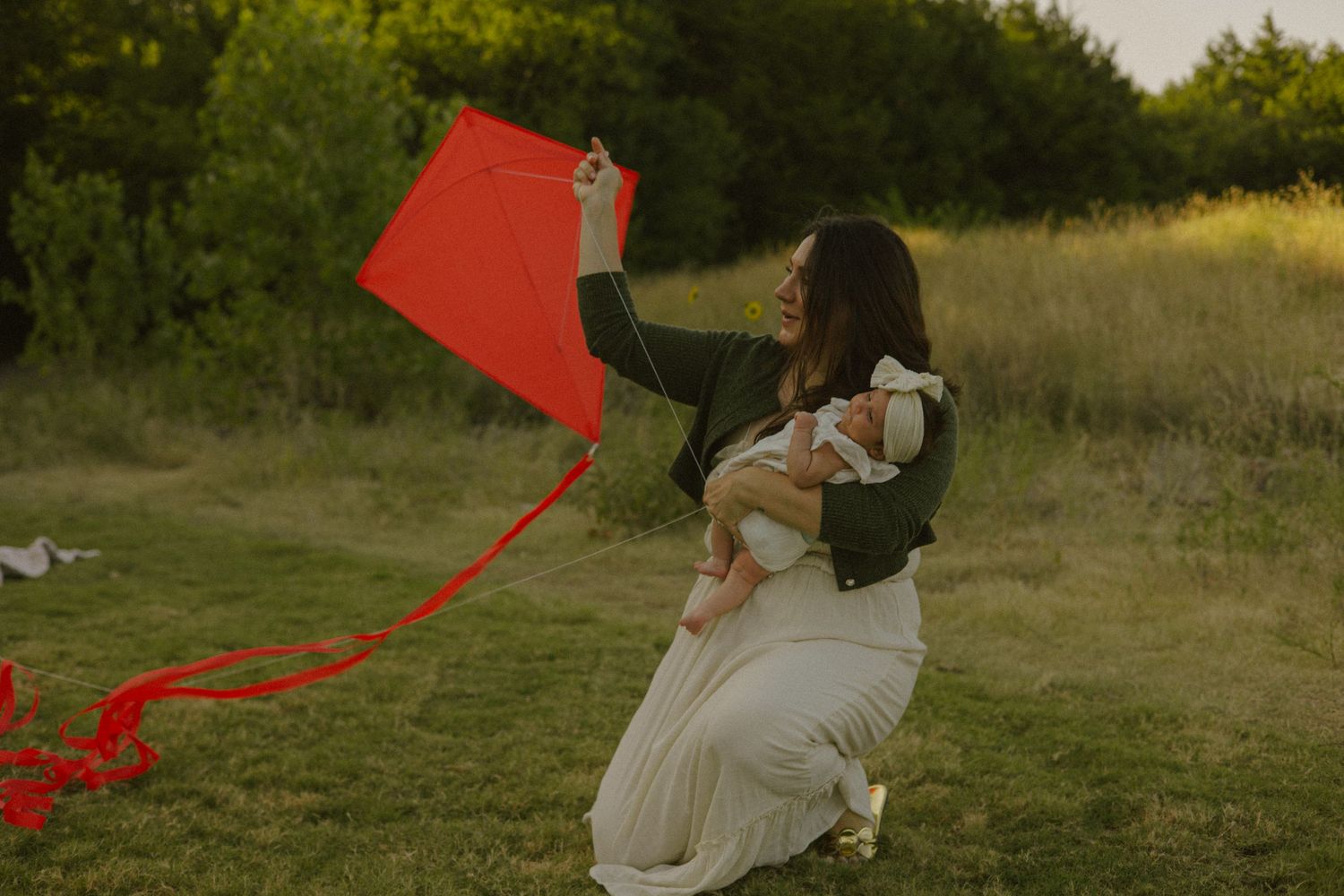 A woman holding a baby and a red kite in a grassy field at golden hour.
