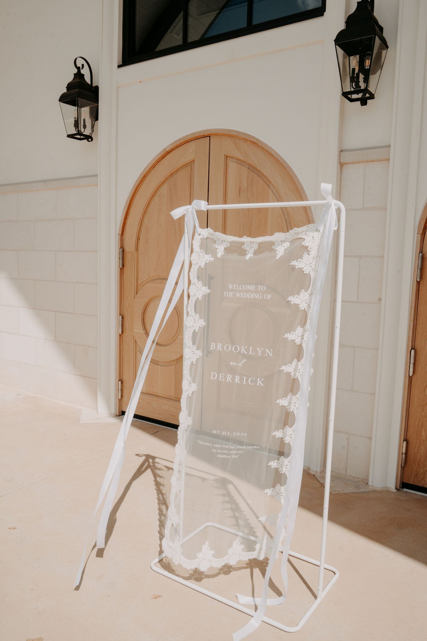 Acrylic wedding welcome sign for Brooklyn and Derrick with lace trim and ribbons on a white frame outside arched wooden doors.