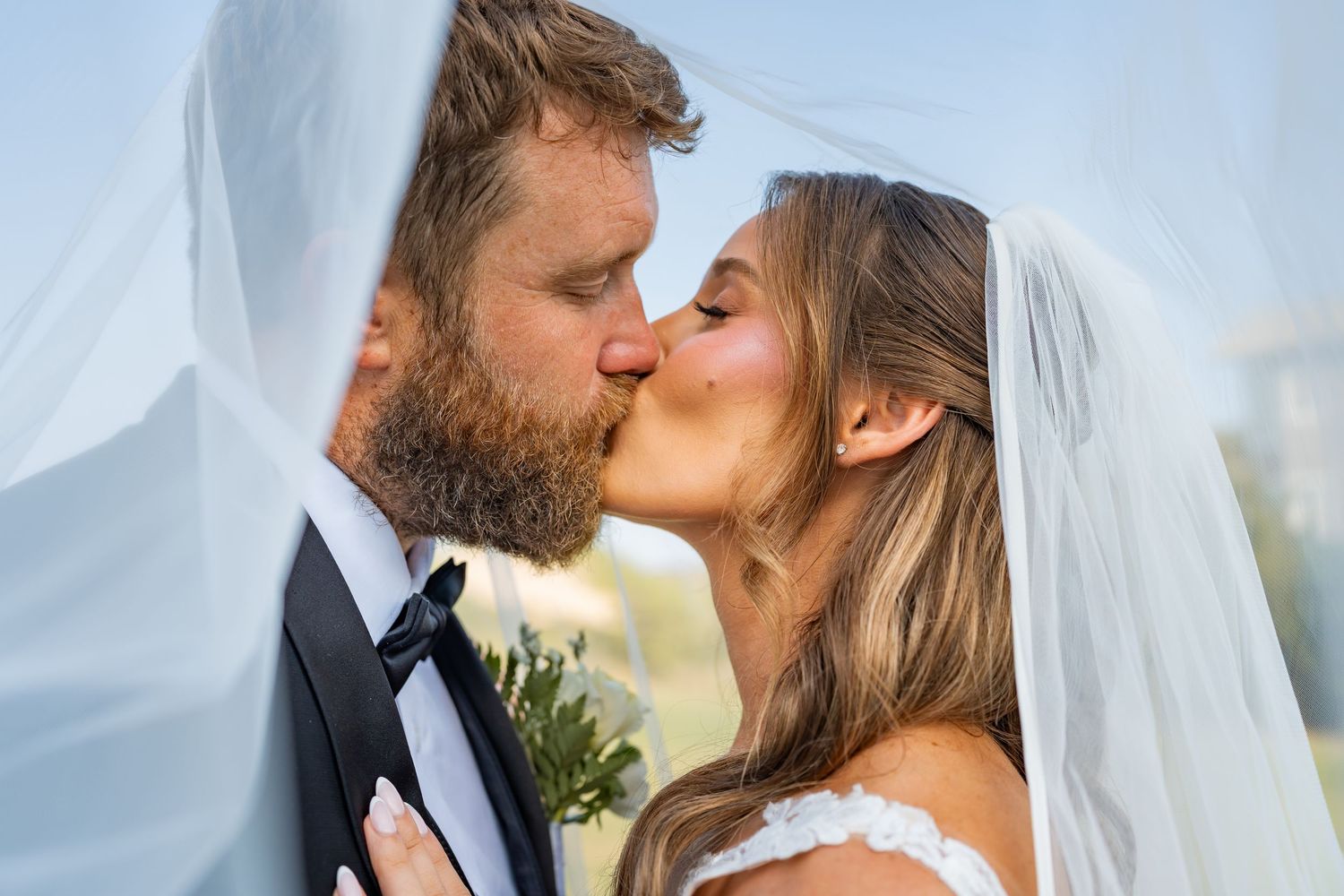 Bride and groom sharing a romantic kiss under a flowing white veil on their wedding day.