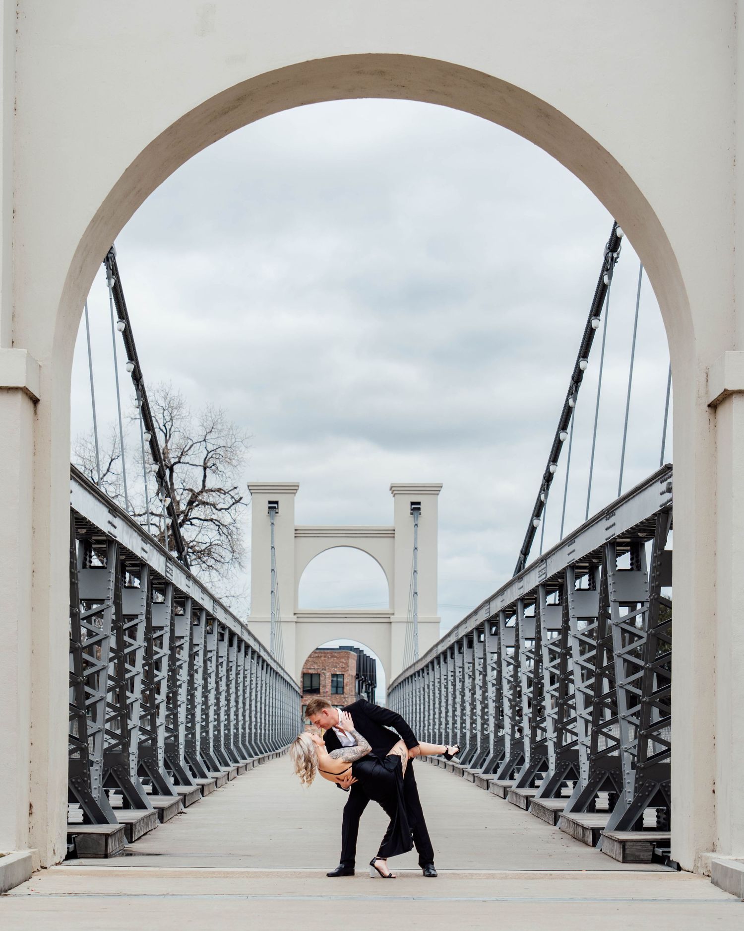 Couple dancing a romantic dip pose on a historic suspension bridge with arched white towers under a cloudy sky.