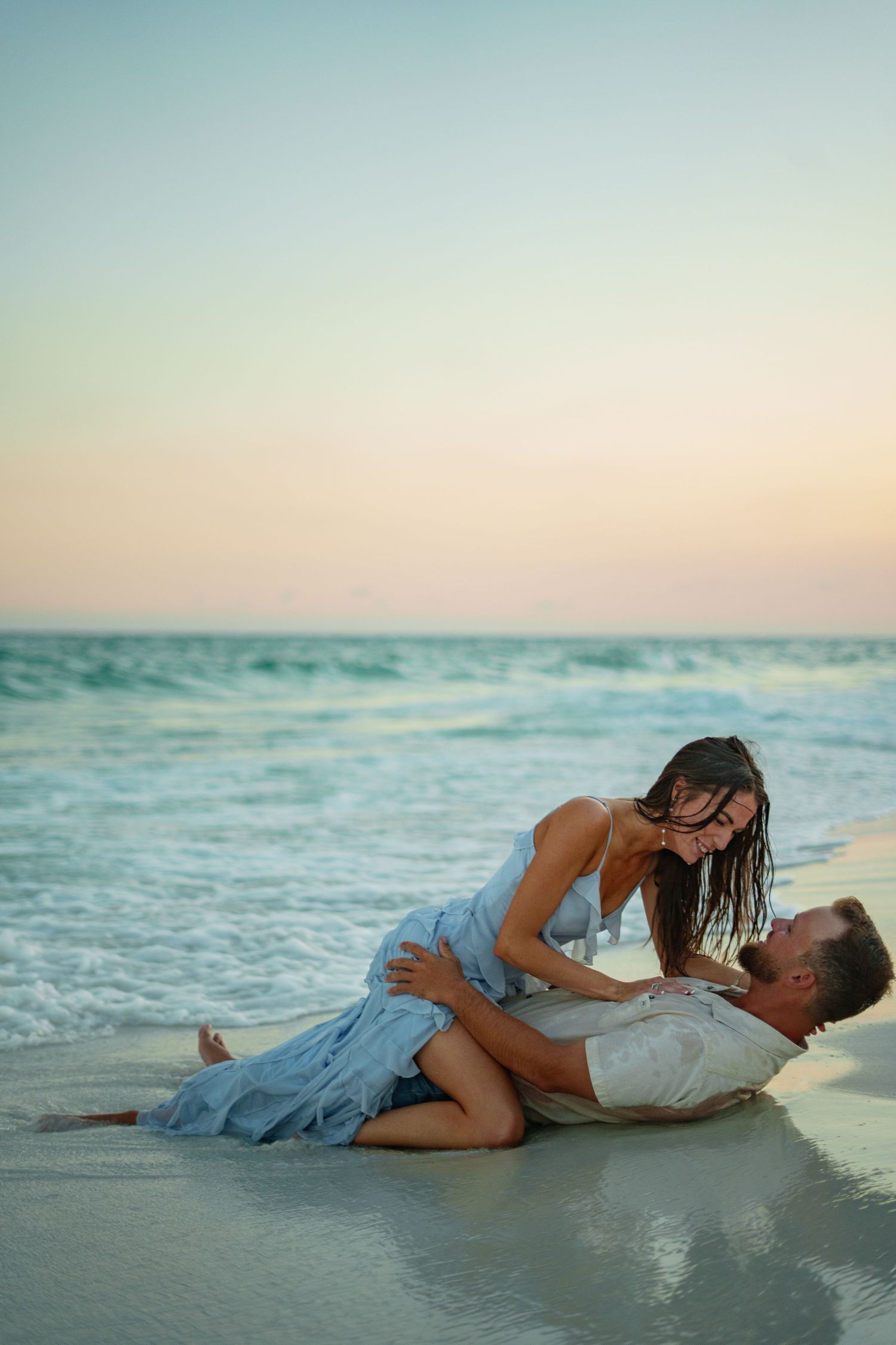 Romantic couple lying together on a sandy beach at sunset, with gentle ocean waves in the background.