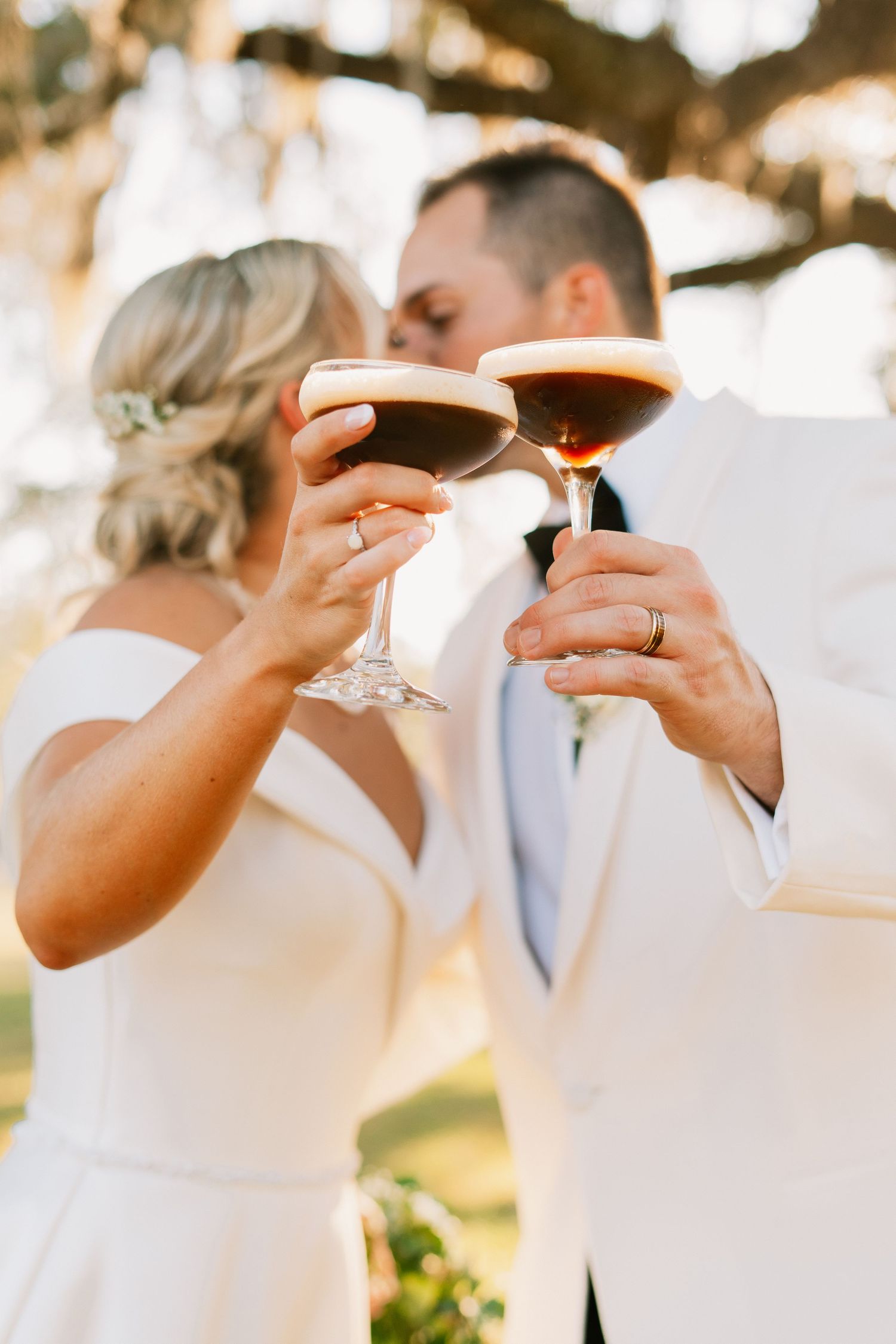 Bride and groom in white attire toasting with espresso martinis during outdoor wedding celebration.