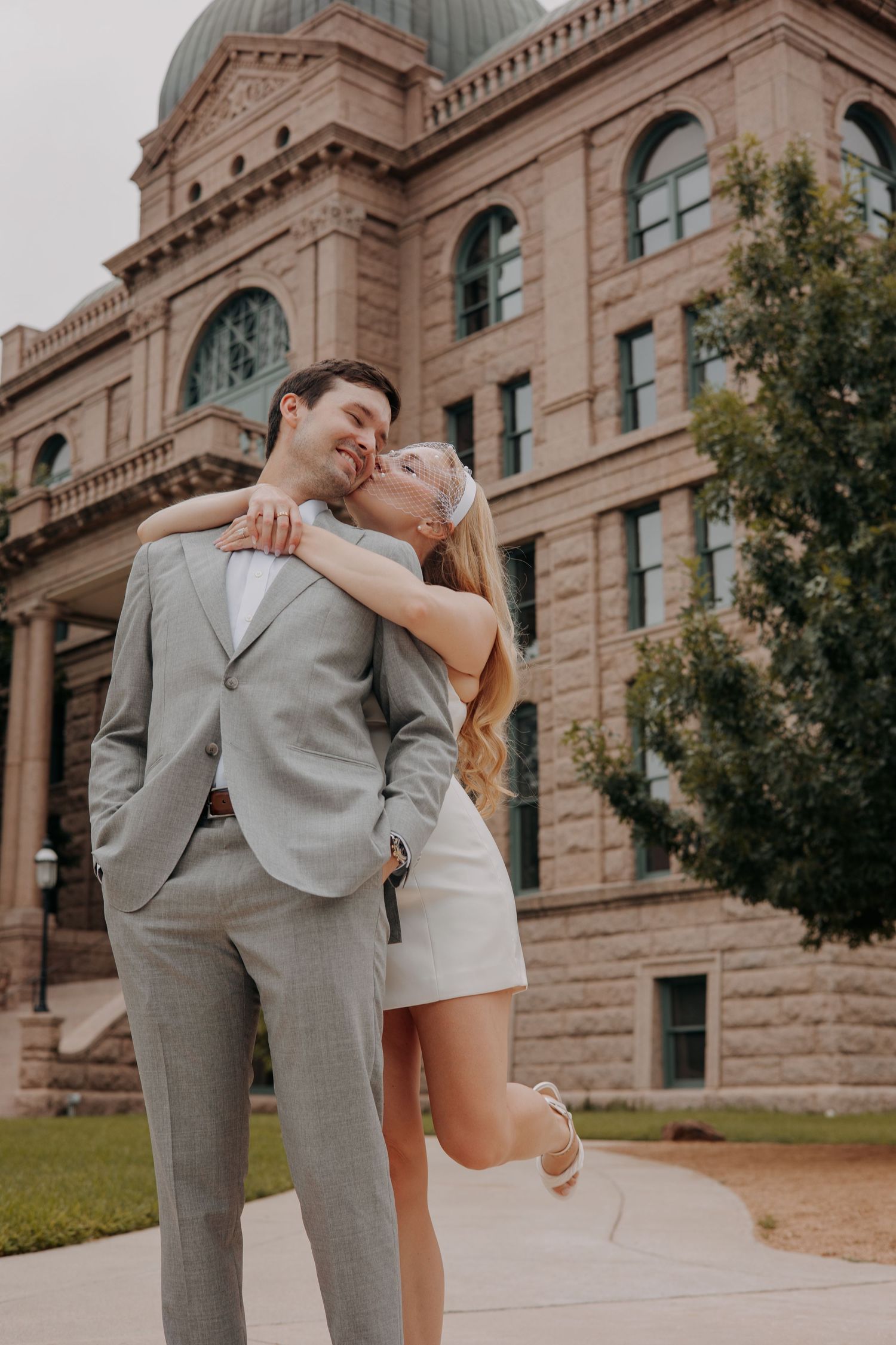 A couple shares a kiss outside a historic stone courthouse, the man in a gray suit and the woman in a white dress kicking up her heel.