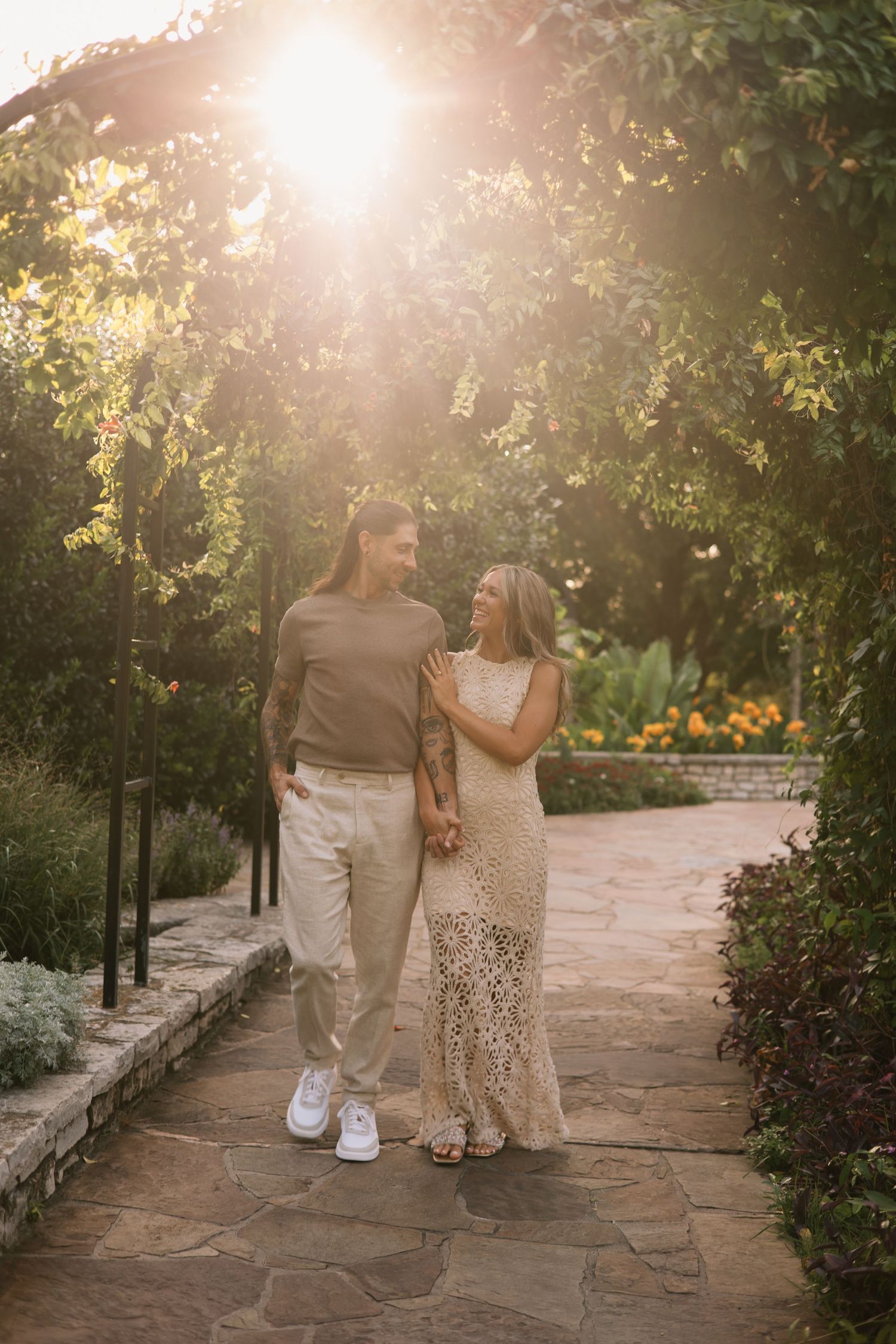 Couple in casual neutral outfits walk hand in hand along a stone garden path under a leafy arbor as golden sunlight filters through.