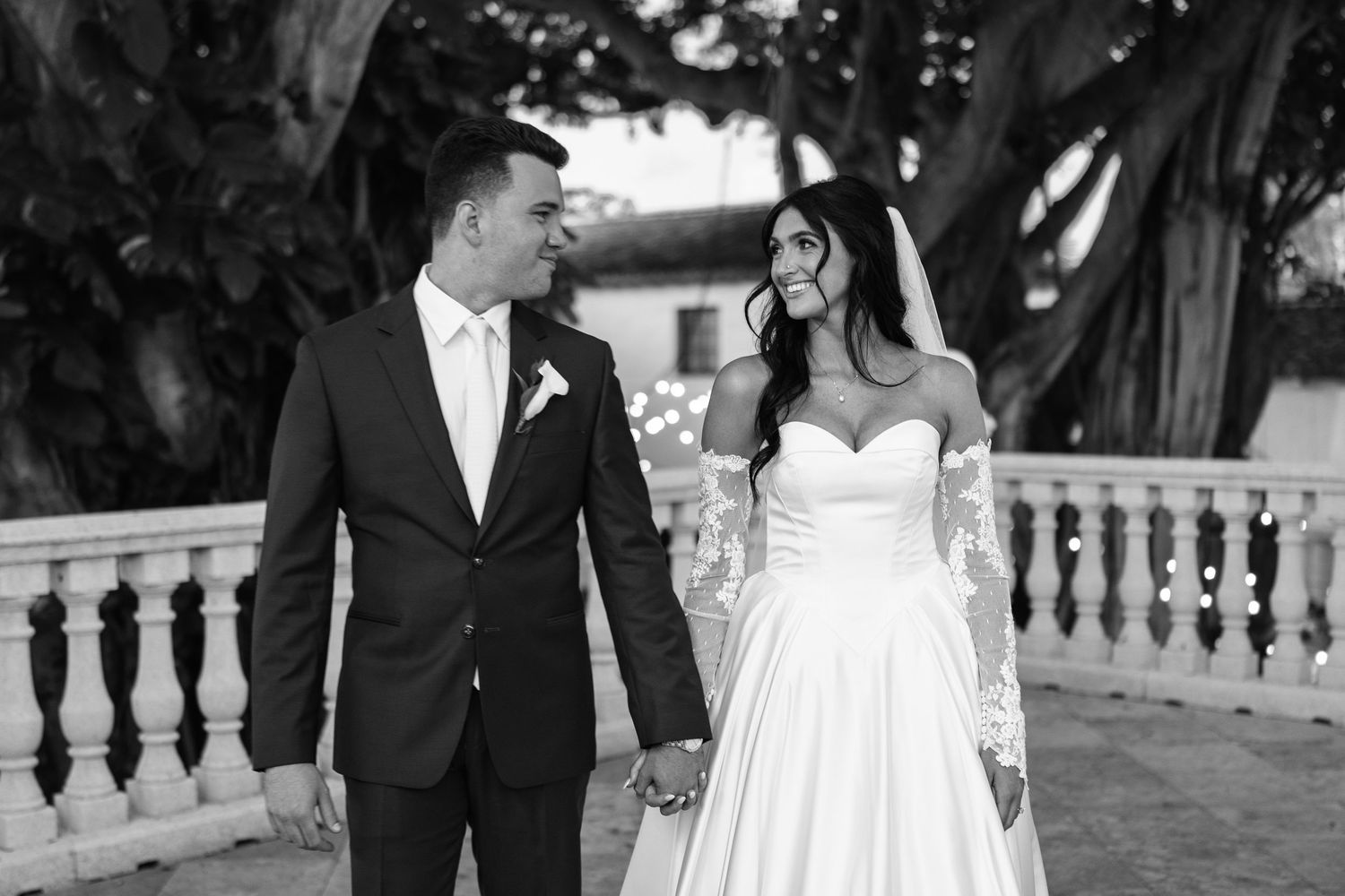 Black and white photo of a bride and groom holding hands outdoors, bride in strapless gown with lace sleeves, groom in dark suit.