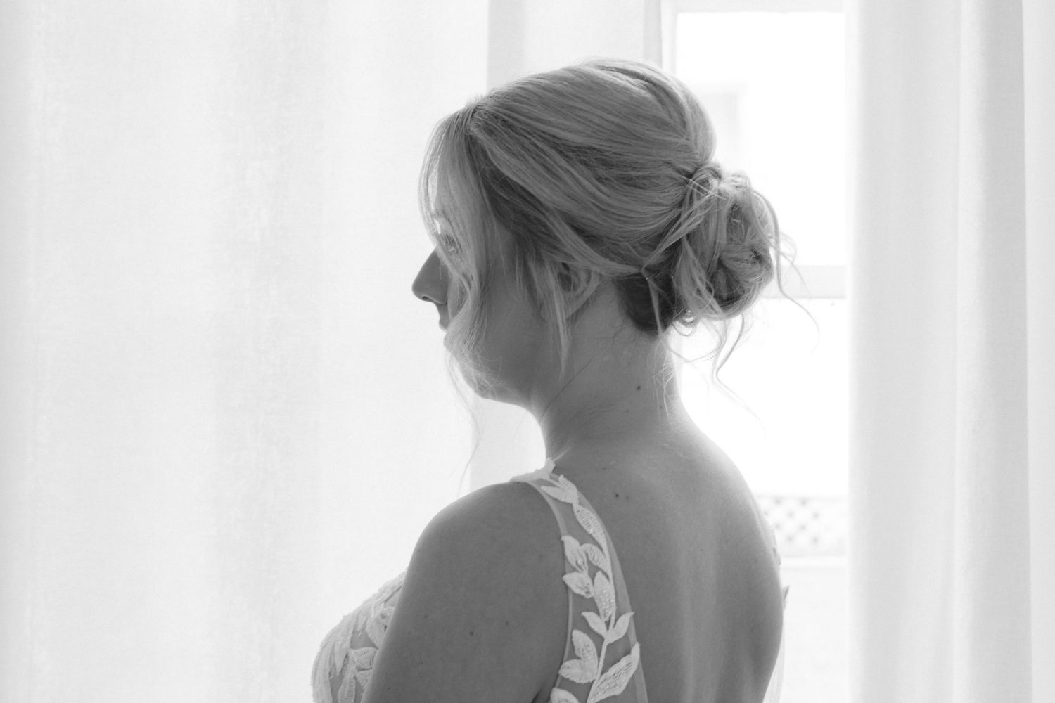 Black and white photo of a bride in a lace dress with an elegant updo, gazing out a bright window.