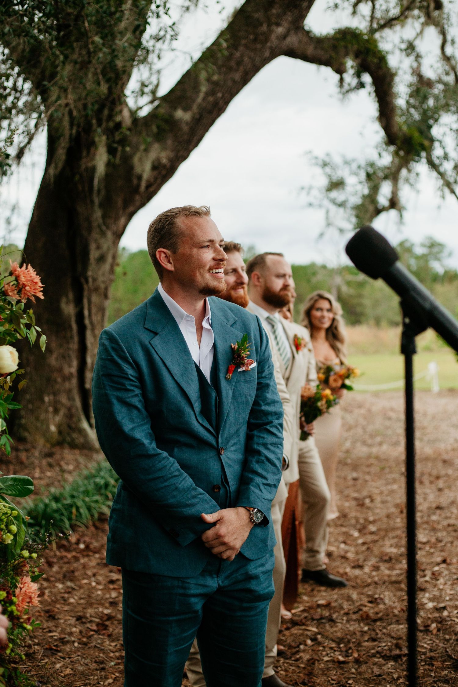 Groom in teal suit smiling at altar during outdoor wedding ceremony with wedding party standing behind him.