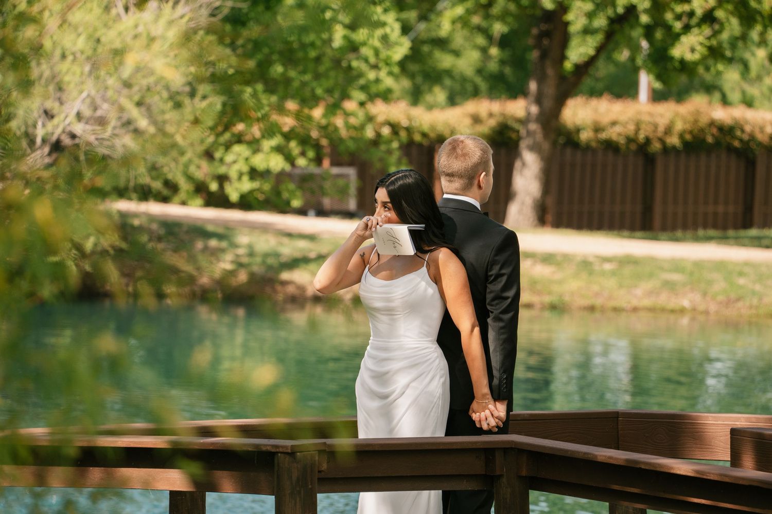 Bride and groom sharing a romantic moment on a wooden bridge overlooking a serene green lake on their wedding day.