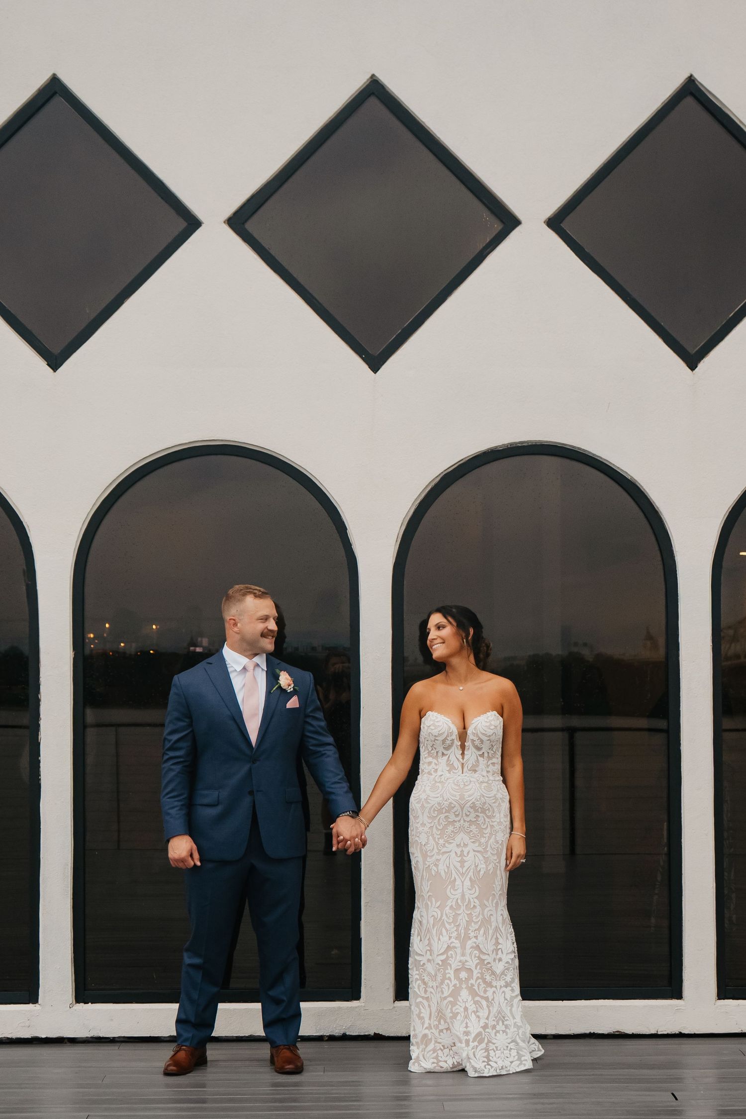 Bride and groom hold hands in front of white arched windows with diamond accents on their wedding day.