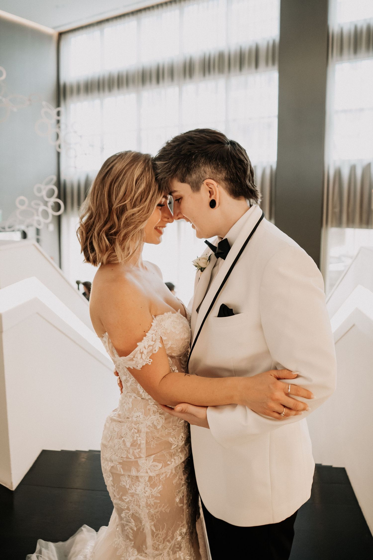 A bride and groom in wedding attire share an intimate moment, touching foreheads and smiling in an elegant indoor venue.