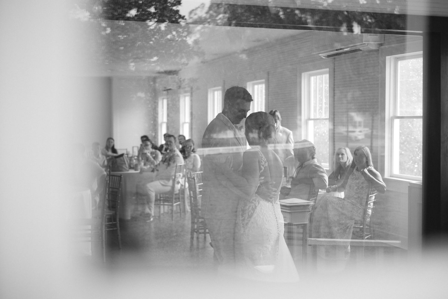 Black and white photo of a couple sharing their first dance at a wedding reception with guests seated in the background.