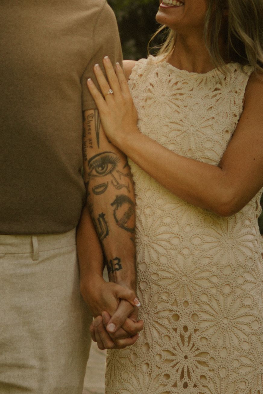 Couple holding hands in a close embrace, a woman in a cream lace dress with a ring beside a man with a tattooed forearm.
