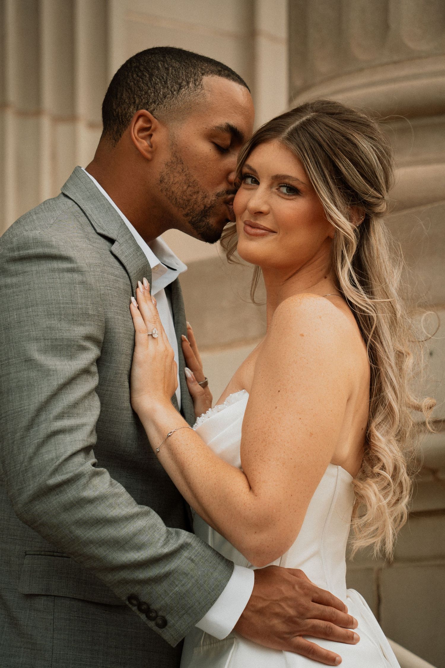 A groom in a gray suit kisses his bride in a white dress on the cheek as she smiles near stone columns.