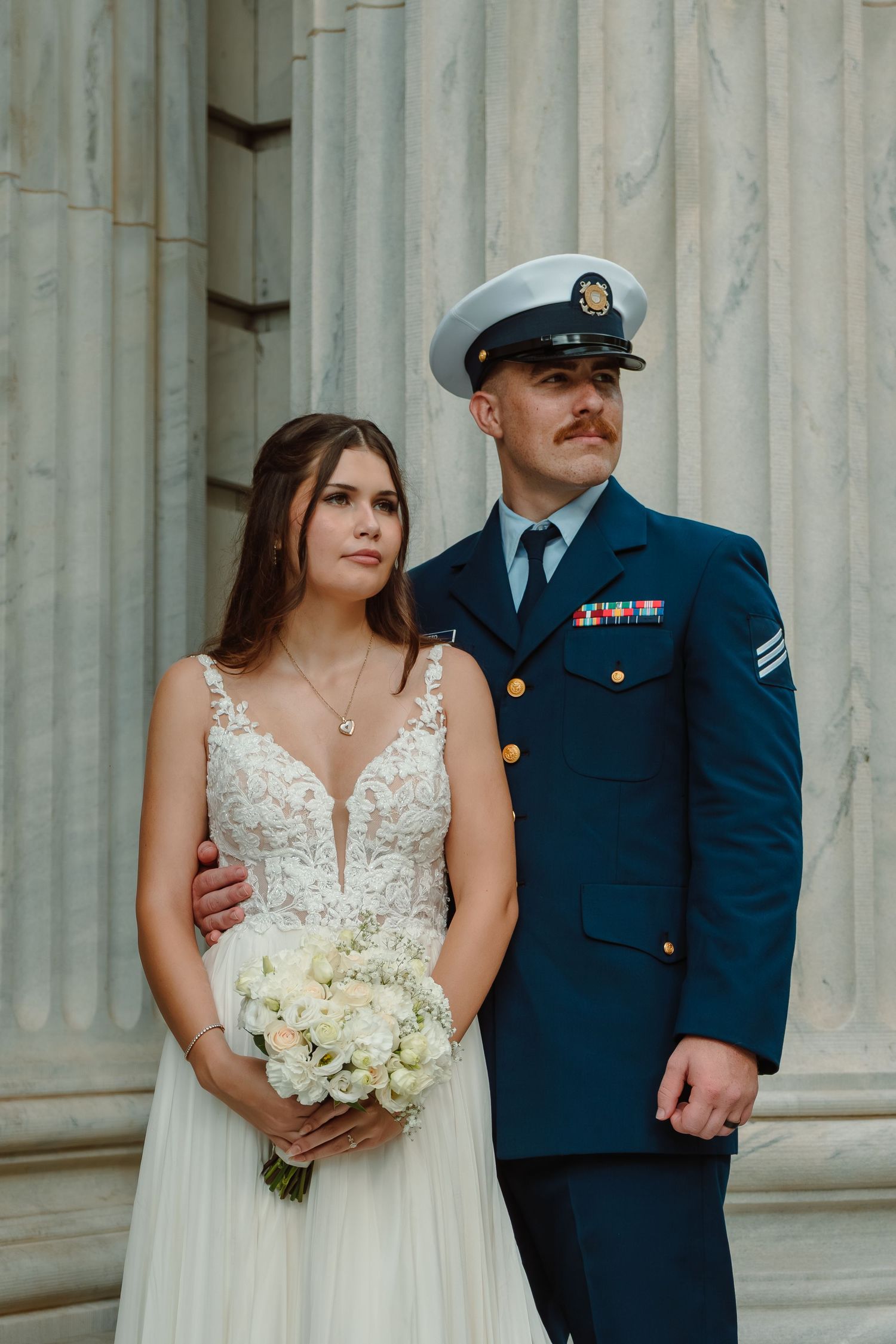Military groom in dress uniform standing proudly with bride holding white bouquet at stone columns.