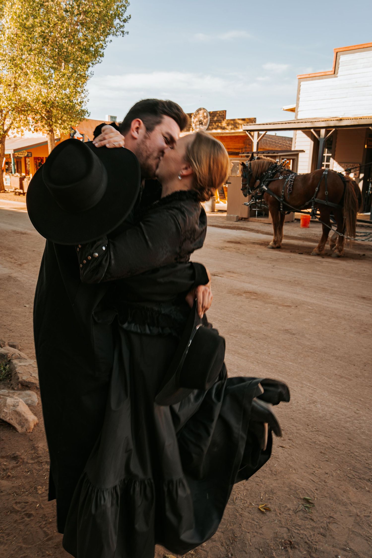 Couple in black Western attire sharing a romantic kiss on a dusty Old West street with horses in the background.