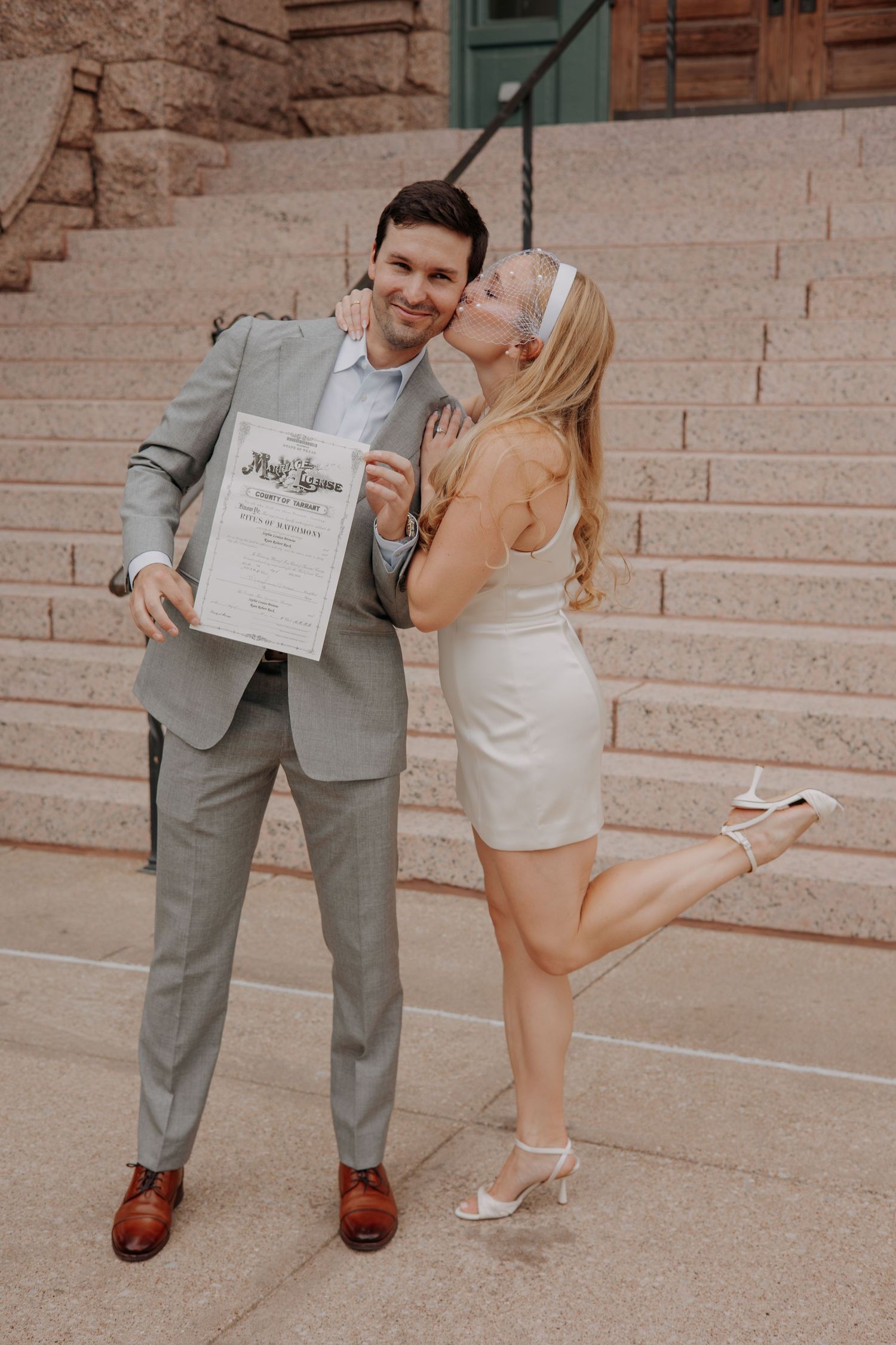 Couple celebrating courthouse wedding on steps, groom holding marriage certificate while bride kisses his cheek, wearing white mini dress.