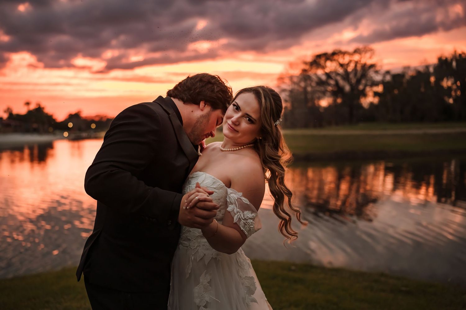 Bride and groom share a romantic embrace at sunset by a lake, with a vivid orange and red sky reflecting on the water.