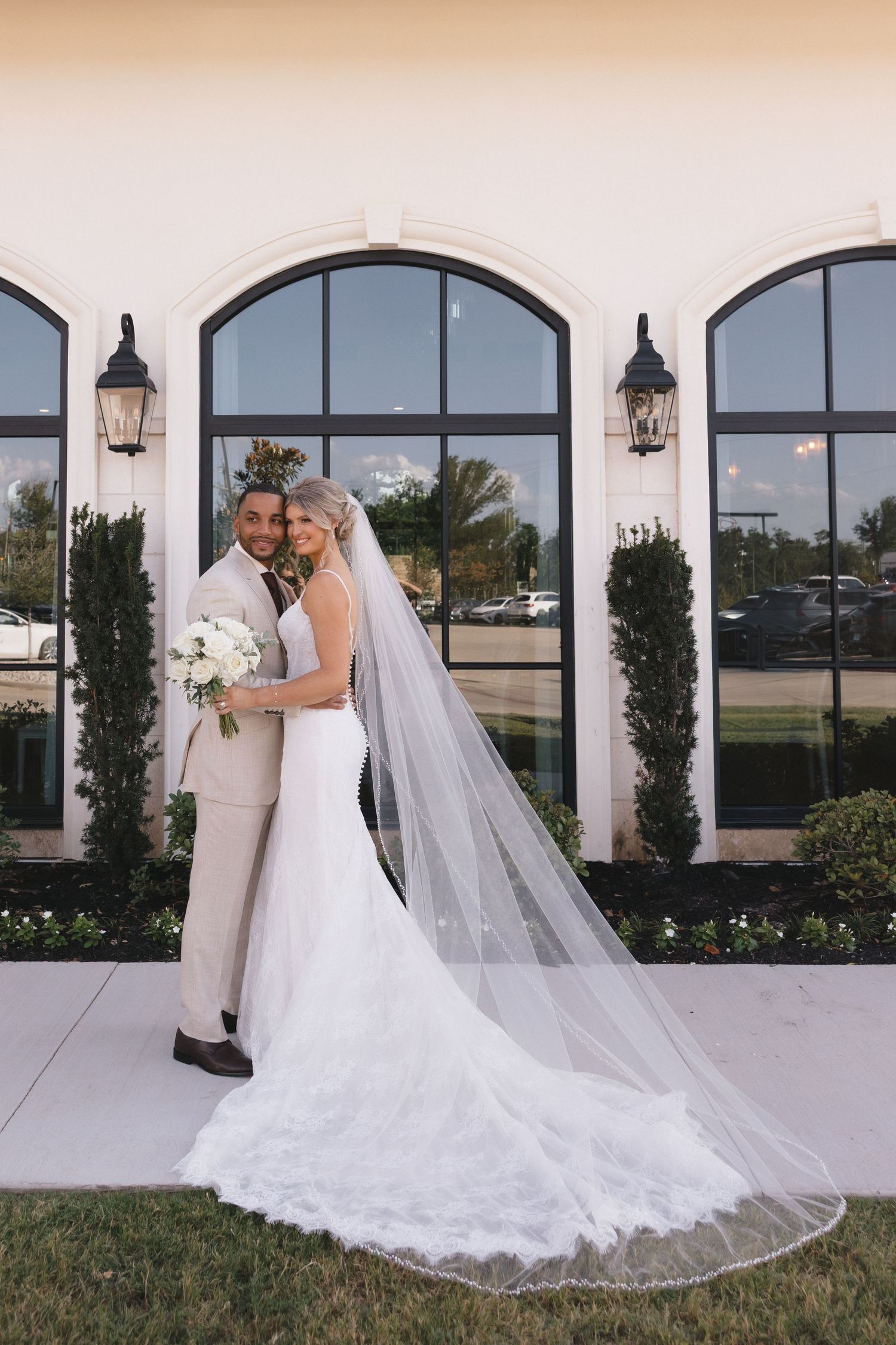 Bride in a lace wedding gown with a long veil embraces her groom in a tan suit while holding a white bouquet outside an arched-window venue.