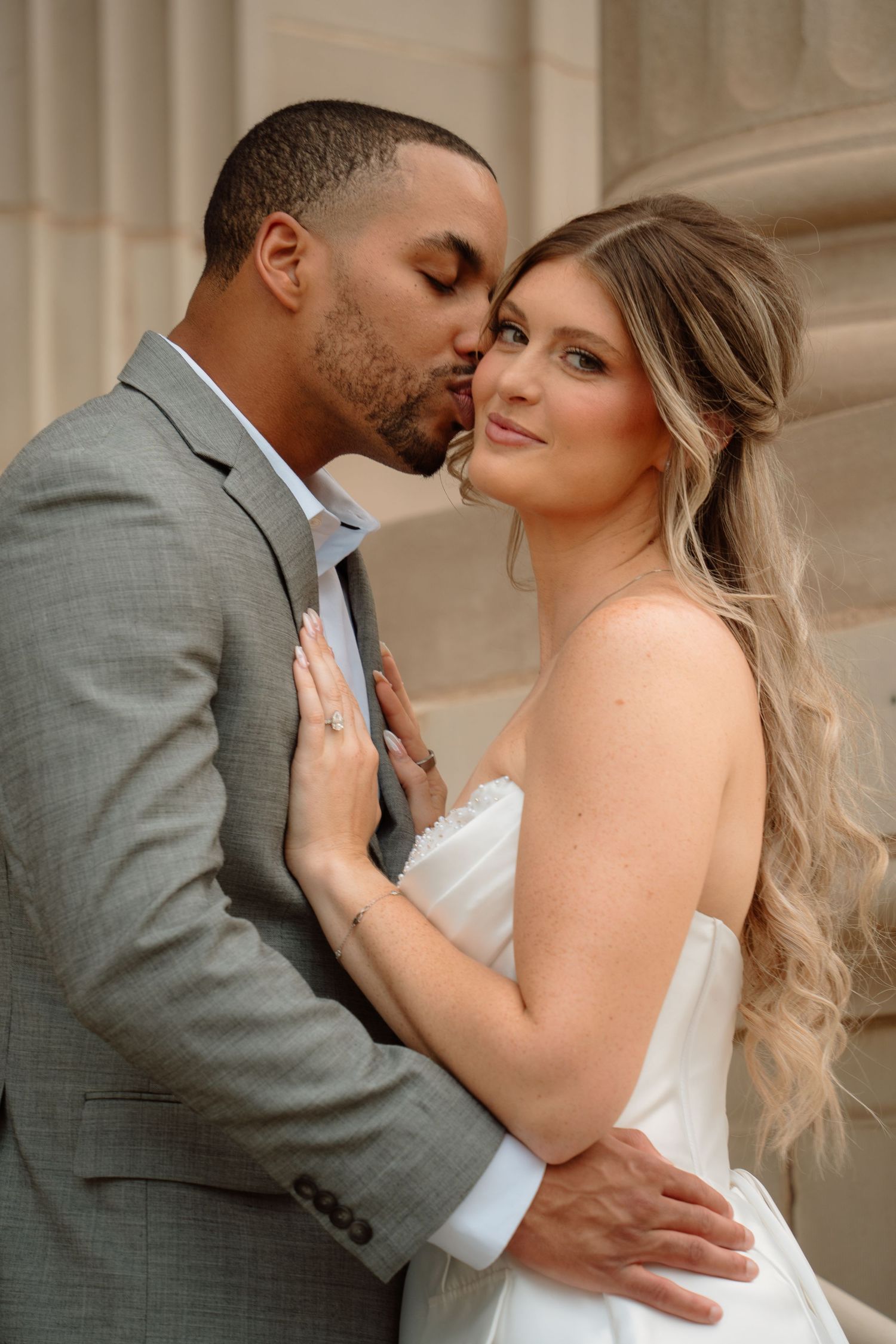 A man in a gray suit kisses the forehead of a smiling woman in a white strapless dress while embracing her near stone columns.