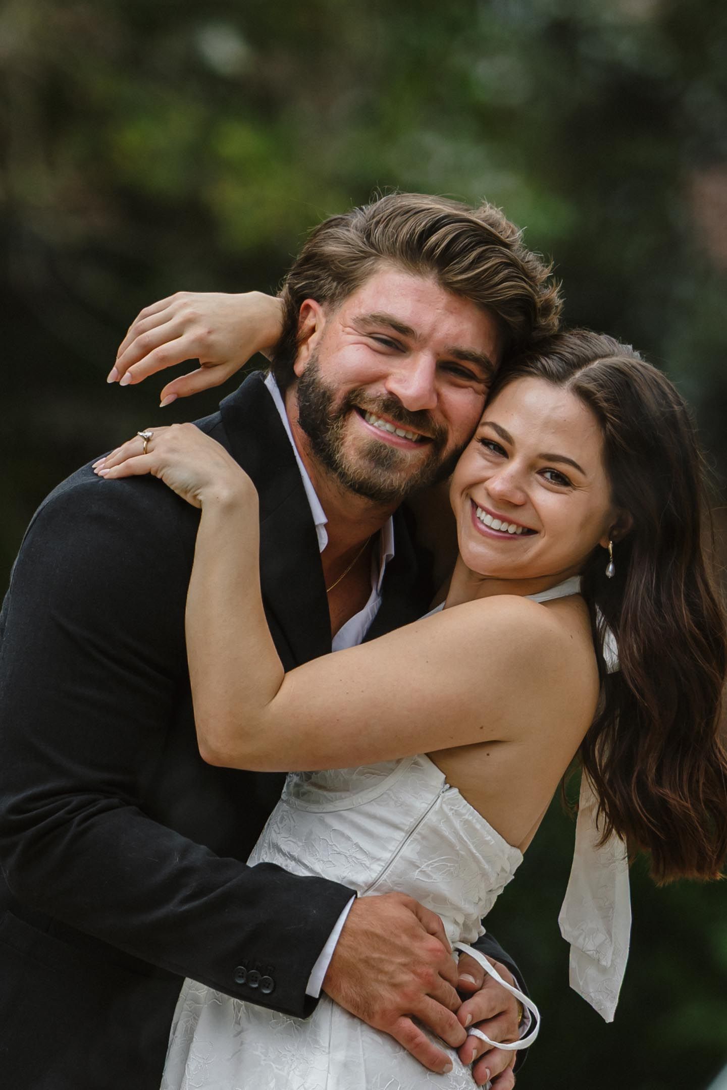 Smiling bearded man in a dark blazer embraces a joyful woman in a white dress outdoors, with a softly blurred green background.