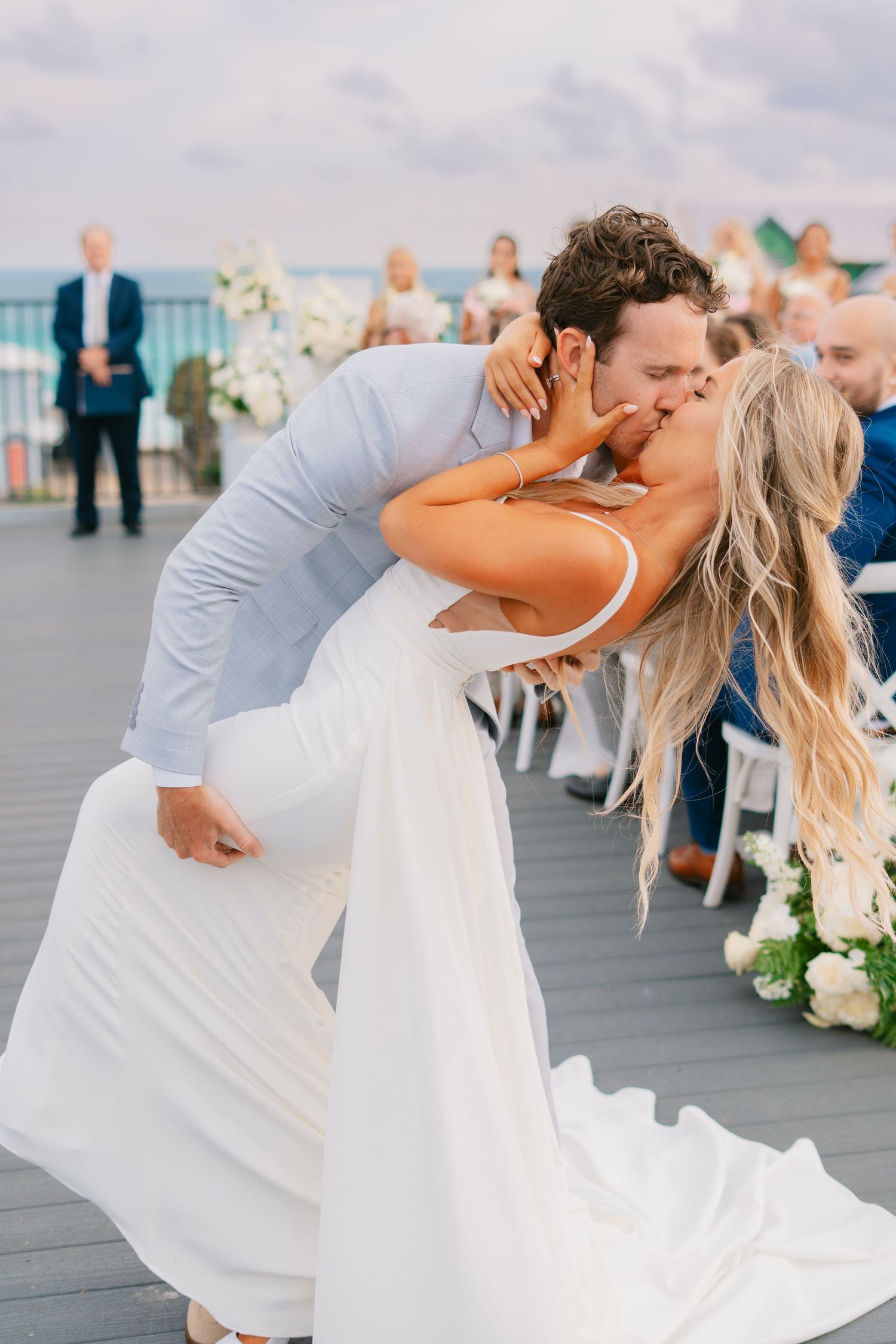 Groom dips bride for a romantic kiss at outdoor wedding ceremony on a deck with white floral arrangements.