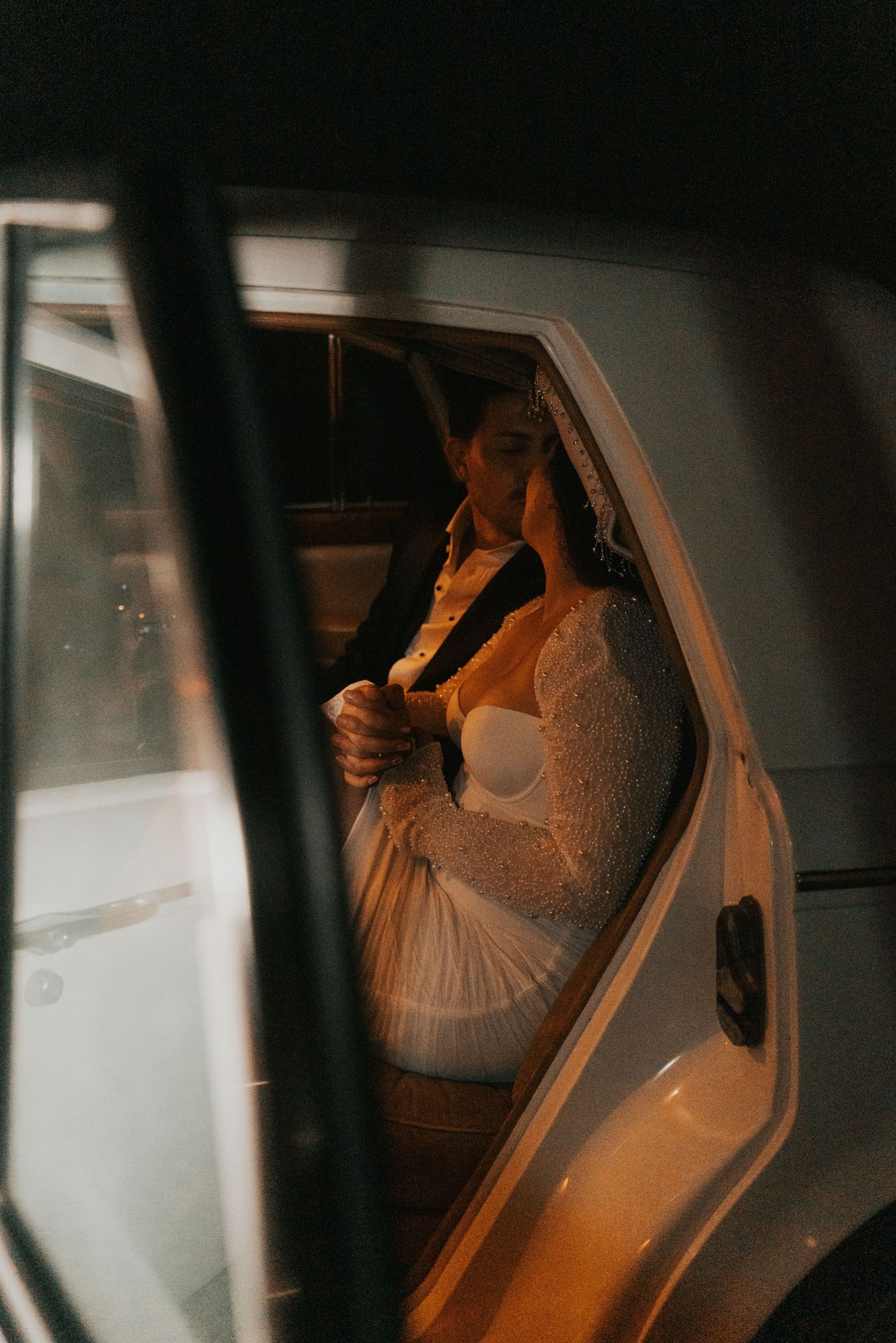 Bride and groom sharing an intimate moment inside a white car at night, bride wearing a lace wedding dress.