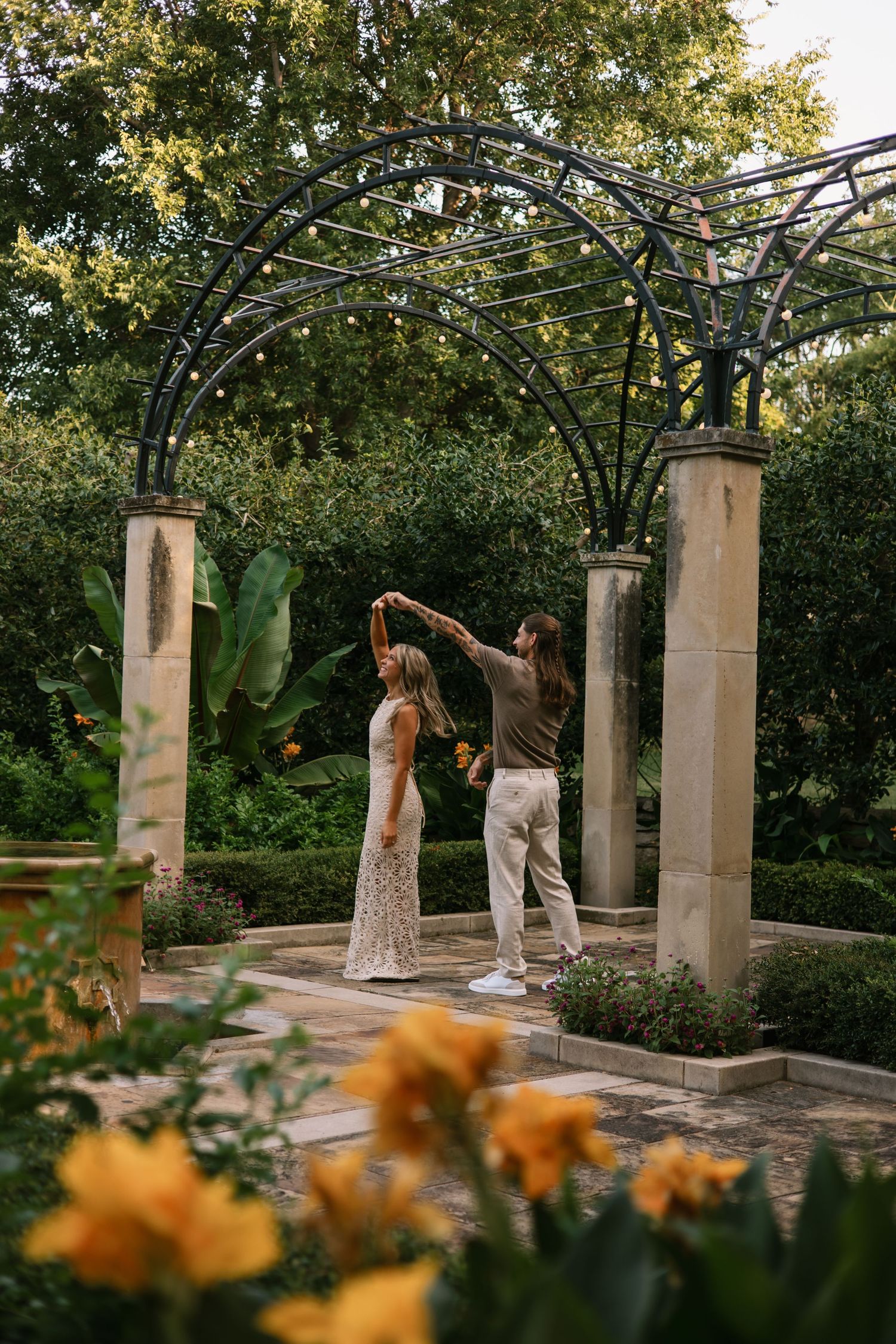 Couple dancing under iron arch pergola in lush garden with string lights and yellow flowers.