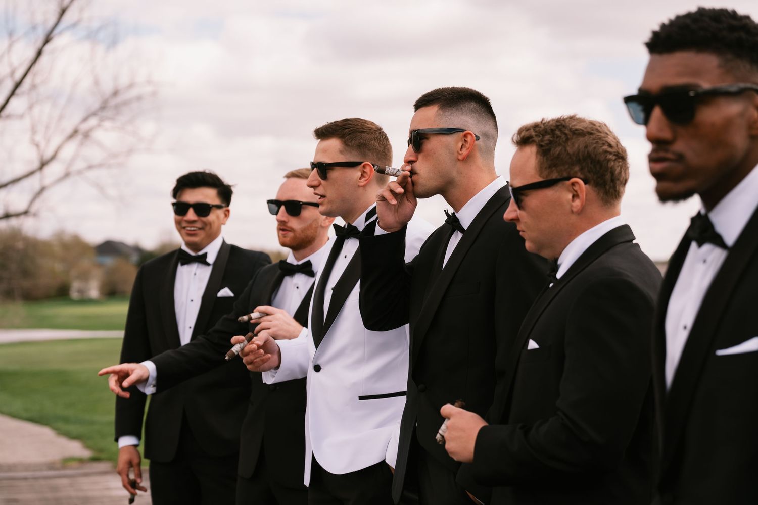 Groomsmen in black tuxedos and sunglasses stand outdoors, with groom in white jacket, at a wedding.
