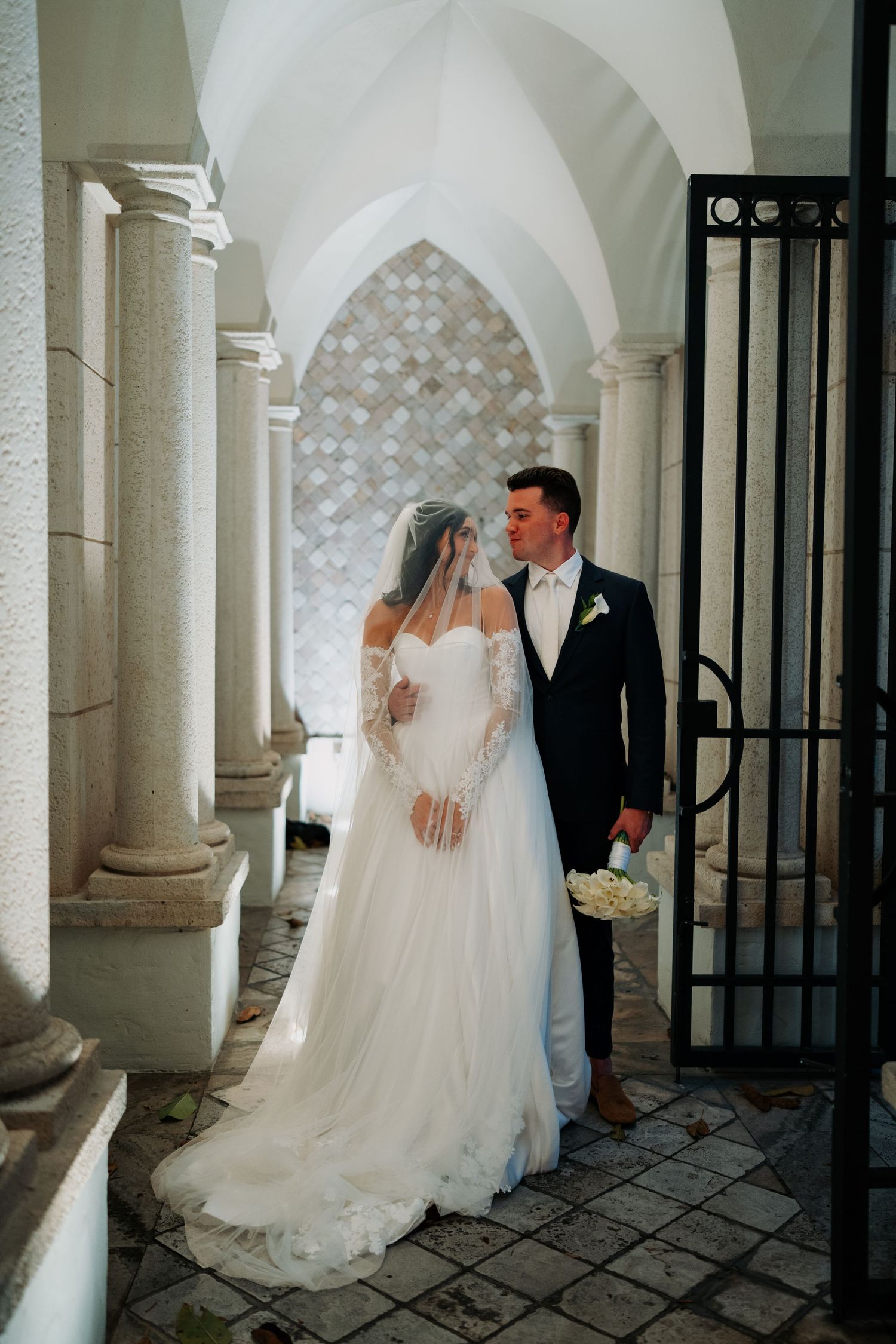 Bride and groom share a romantic moment under arched columns at an elegant wedding venue.