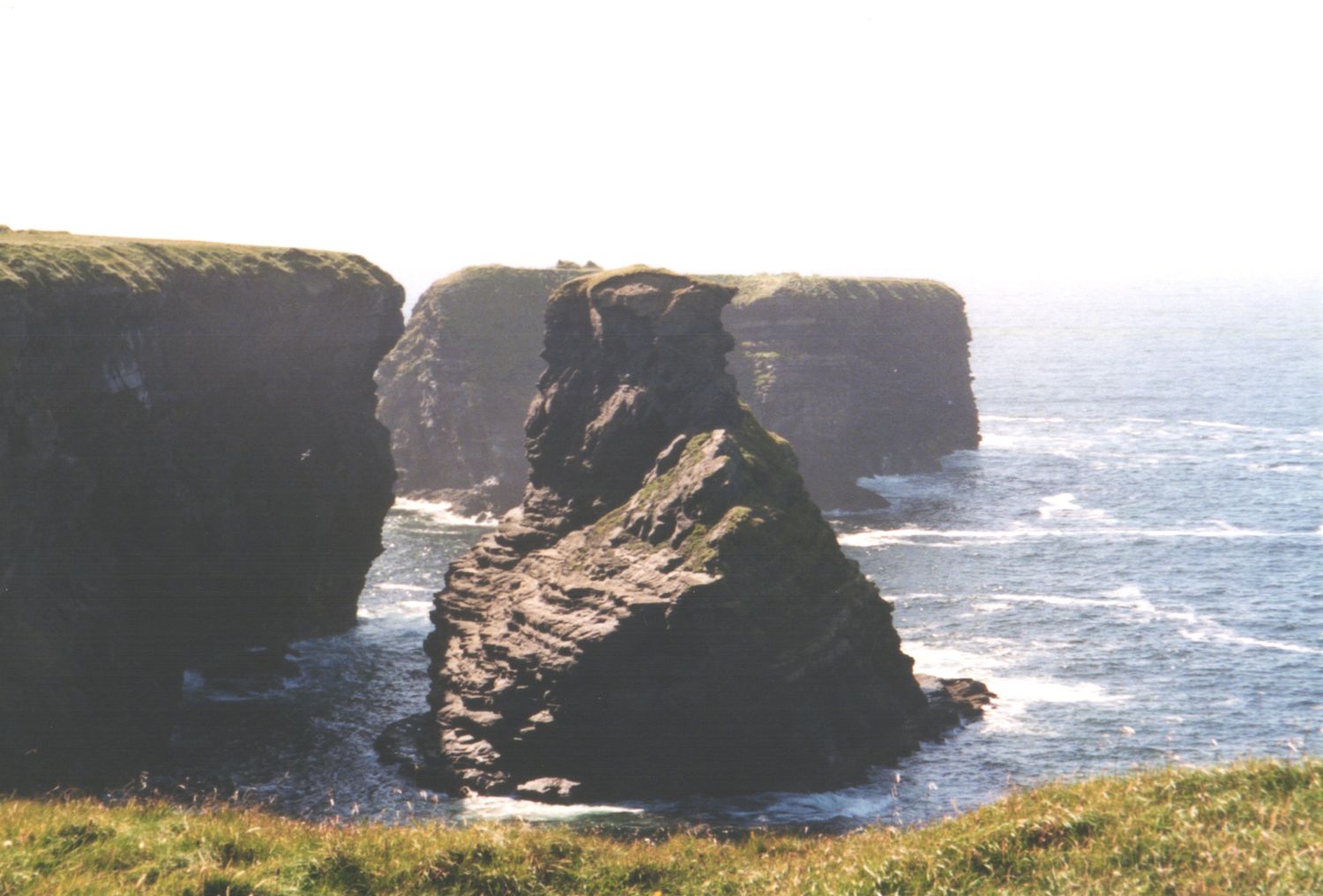 Dramatic rocky sea stacks rise from crashing ocean waves along a rugged coastal cliff landscape.