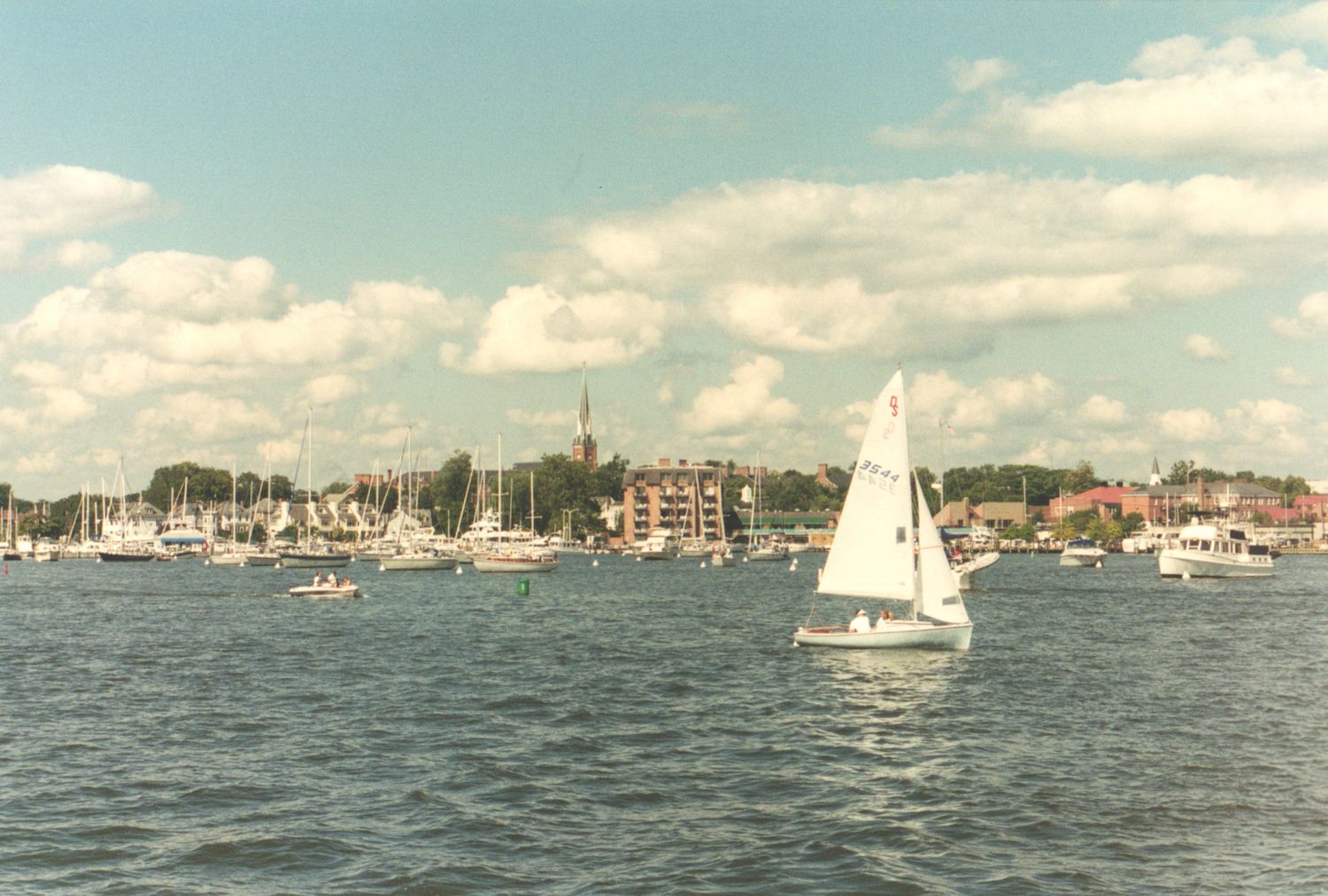Sailboats gliding across a calm harbor with a picturesque waterfront town and cloudy blue sky in the background.