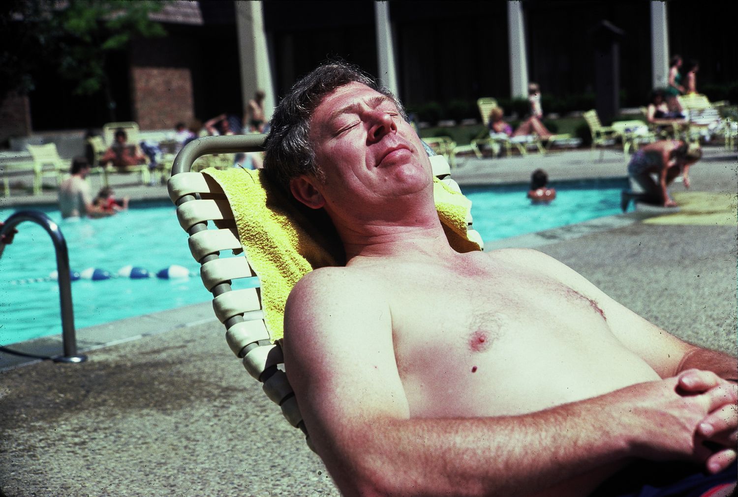 Shirtless man relaxing on lounge chair sunbathing by outdoor swimming pool on sunny day.
