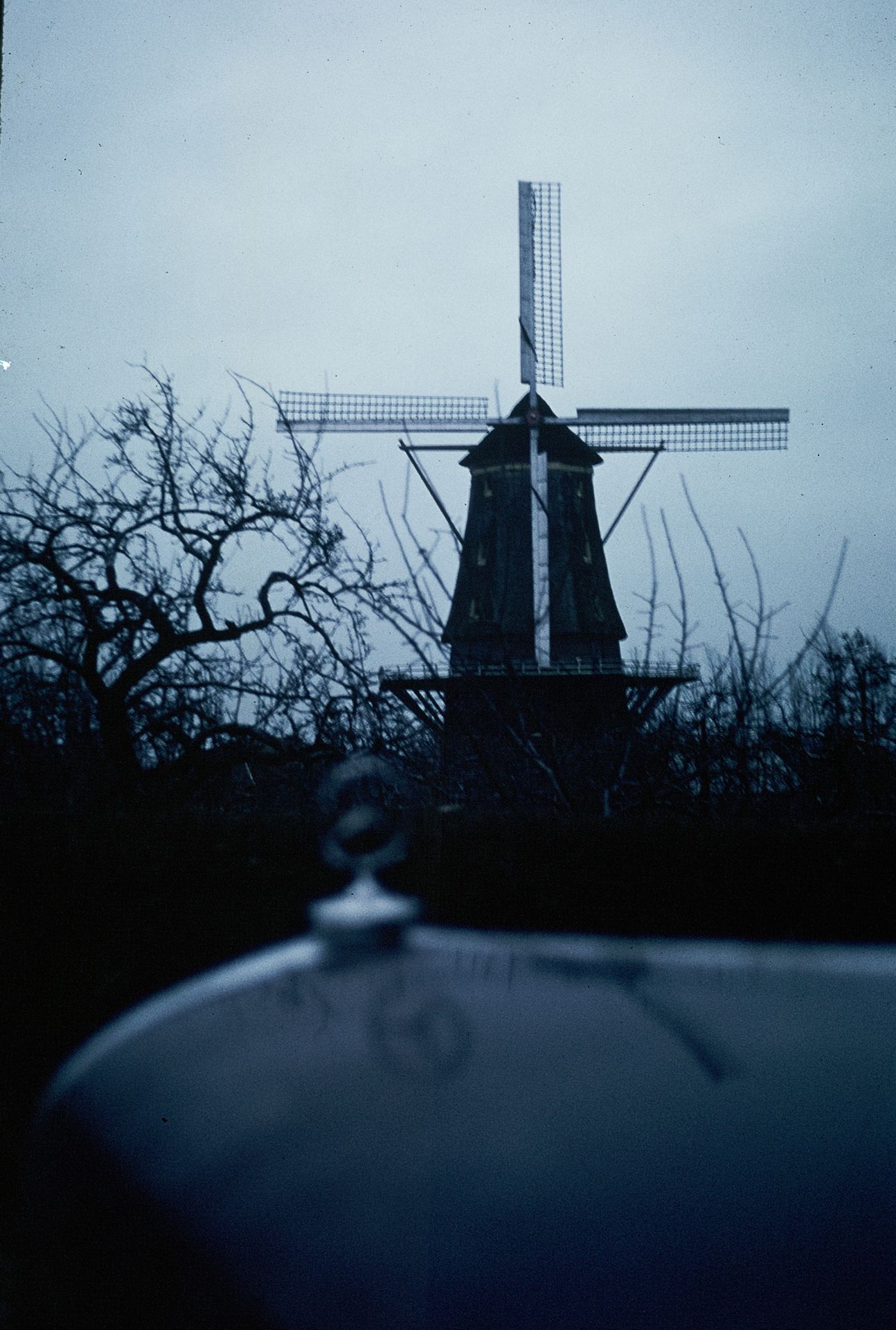 Silhouette of a traditional Dutch windmill at dusk with bare trees and car hood ornament.